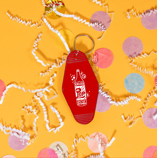On a sunny mustard background sits a keychain with white crinkle and big, colorful confetti scattered around. This vintage, motel keychain is red with a white illustration of a pop bottle and bubbles. It says "IT'S CALLED Pop NOT Soda" in a white, handwritten lettering font.