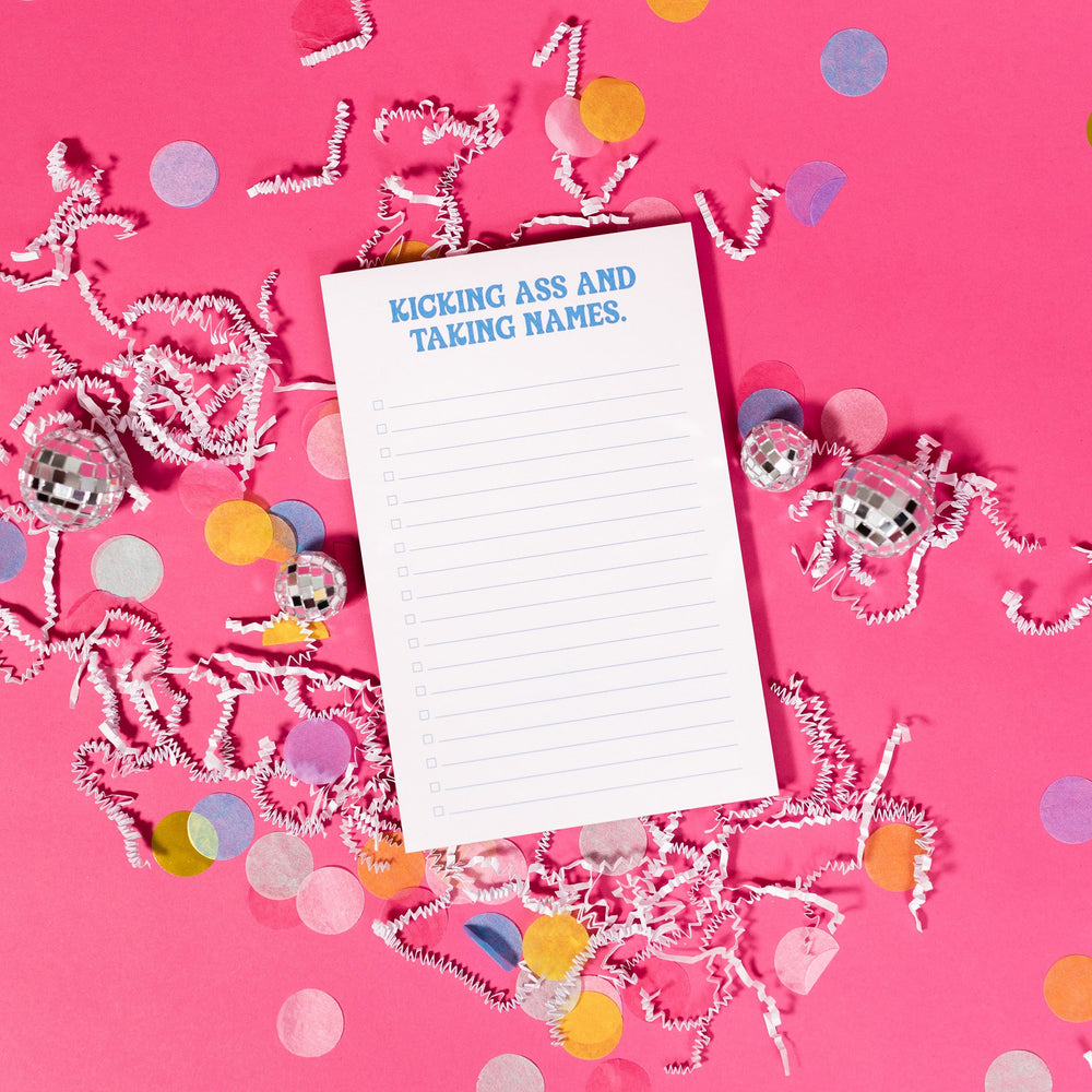 On a hot pink background sits a white notepad with white crinkle and big, colorful confetti scattered around. There are mini disco balls. In a groovy blue lettering it says "KICKING ASS AND TAKING NAMES." There are blue check boxes and lines. 