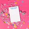 On a hot pink background sits a white notepad with white crinkle and big, colorful confetti scattered around. There are mini disco balls. In a groovy blue lettering it says "KICKING ASS AND TAKING NAMES." There are blue check boxes and lines. 