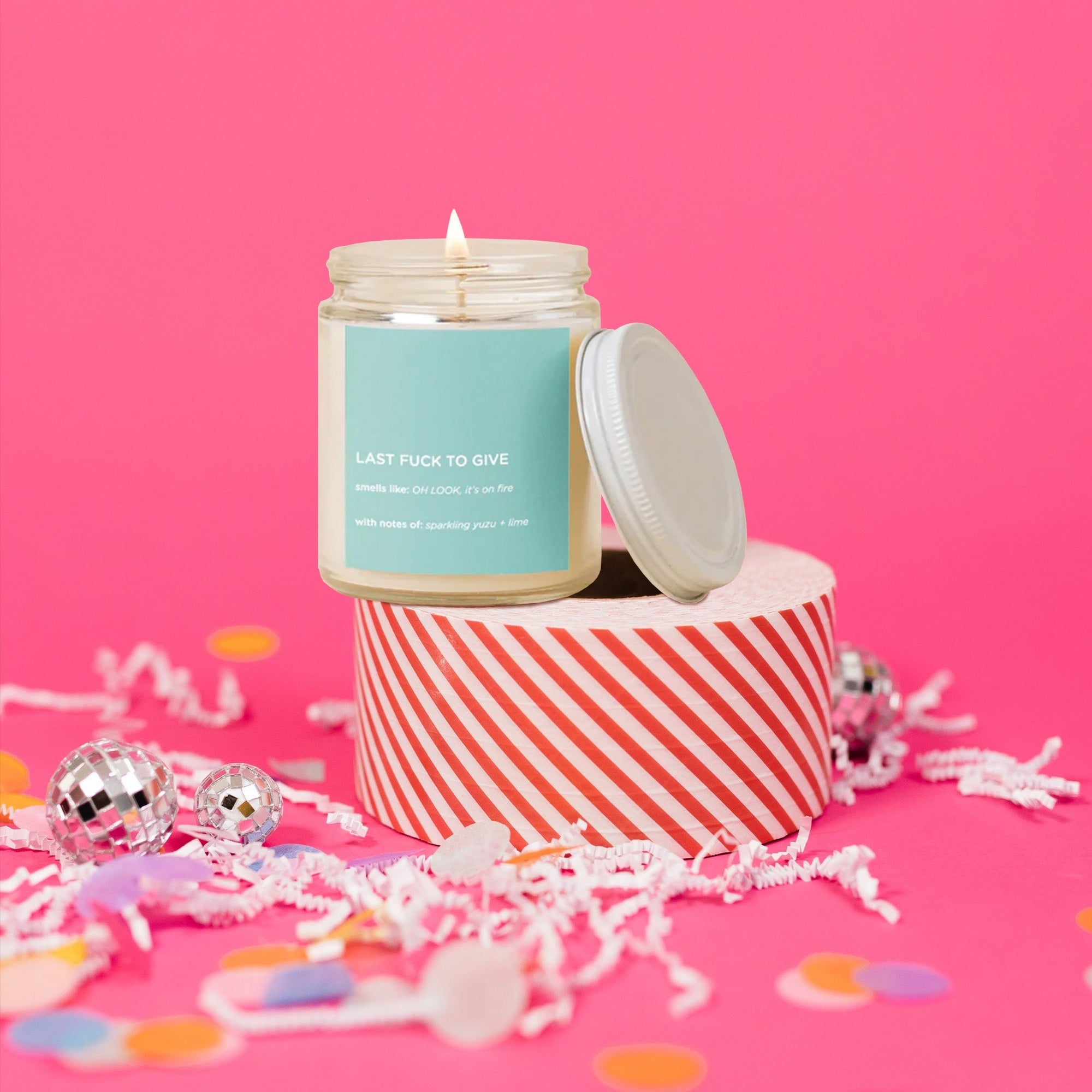 On a hot pink background sits a candle with a lid atop a red and white striped packing tape. There are white crinkle and big, colorful confetti scattered around. The glass candle is lit with a mint label. It says "LAST FUCK TO GIVE" in all caps, white block font. It also says "smells like: OH LOOK, it's on fire," "With notes of: sparkling yuzu + lime." It is in all white, block font. 