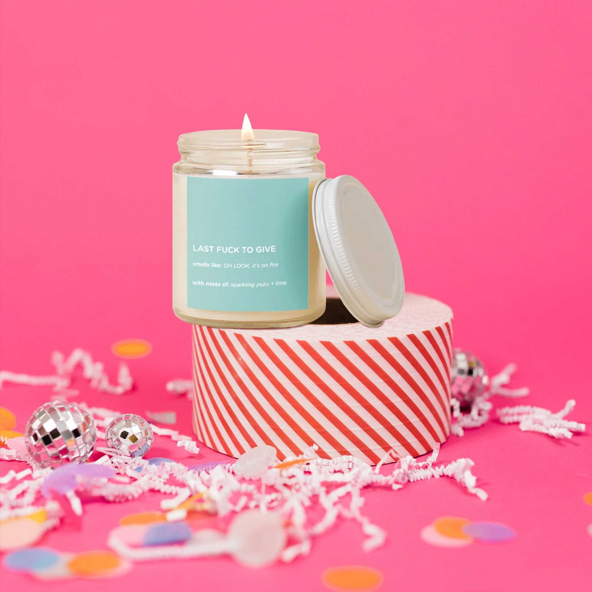 On a hot pink background sits a candle with a lid atop a red and white striped packing tape. There are white crinkle and big, colorful confetti scattered around. The glass candle is lit with a mint label. It says "LAST FUCK TO GIVE" in all caps, white block font. It also says "smells like: OH LOOK, it's on fire," "With notes of: sparkling yuzu + lime." It is in all white, block font. 