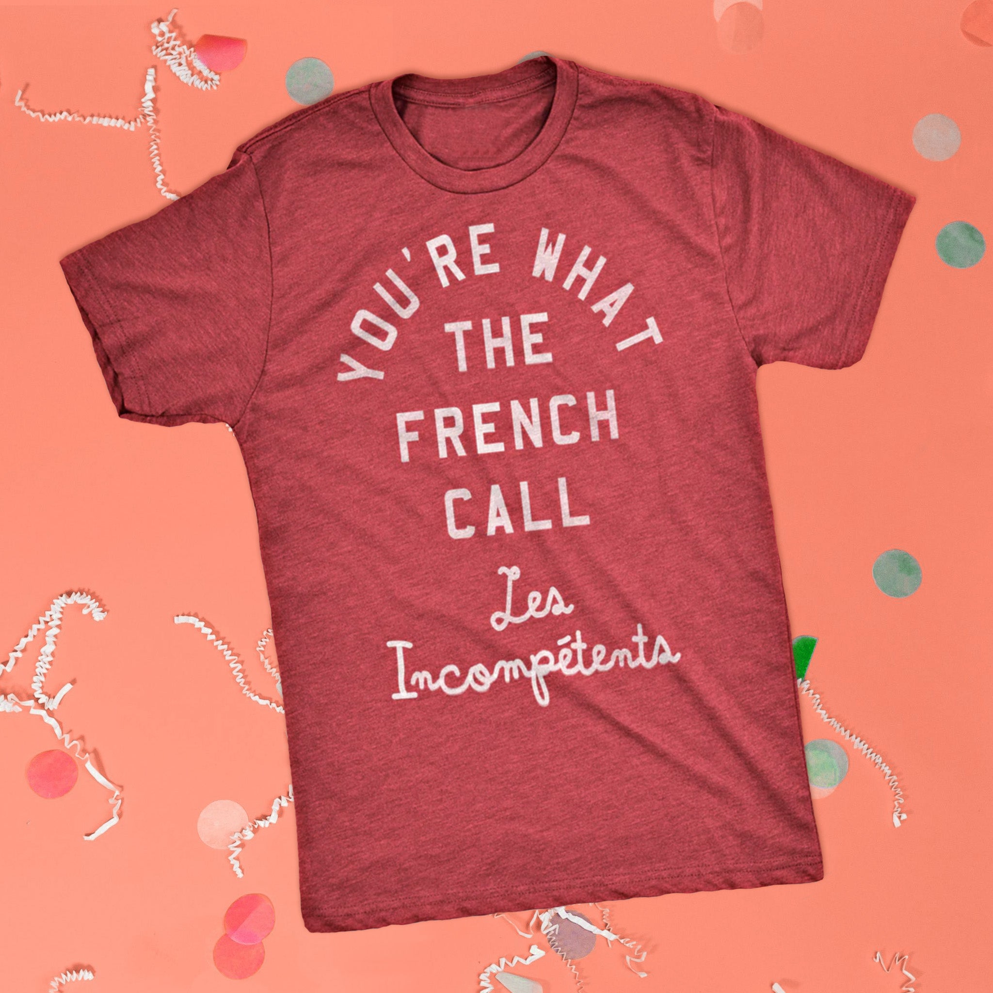 On a pink coral background sits the front of a t-shirt with white crinkle and big, colorful confetti scattered around. This Home Alone Inspired crewneck heathered red t-shirt has white collegiate and script lettering and it says "YOU'RE WHAT THE FRENCH CALL Les Incompetents."