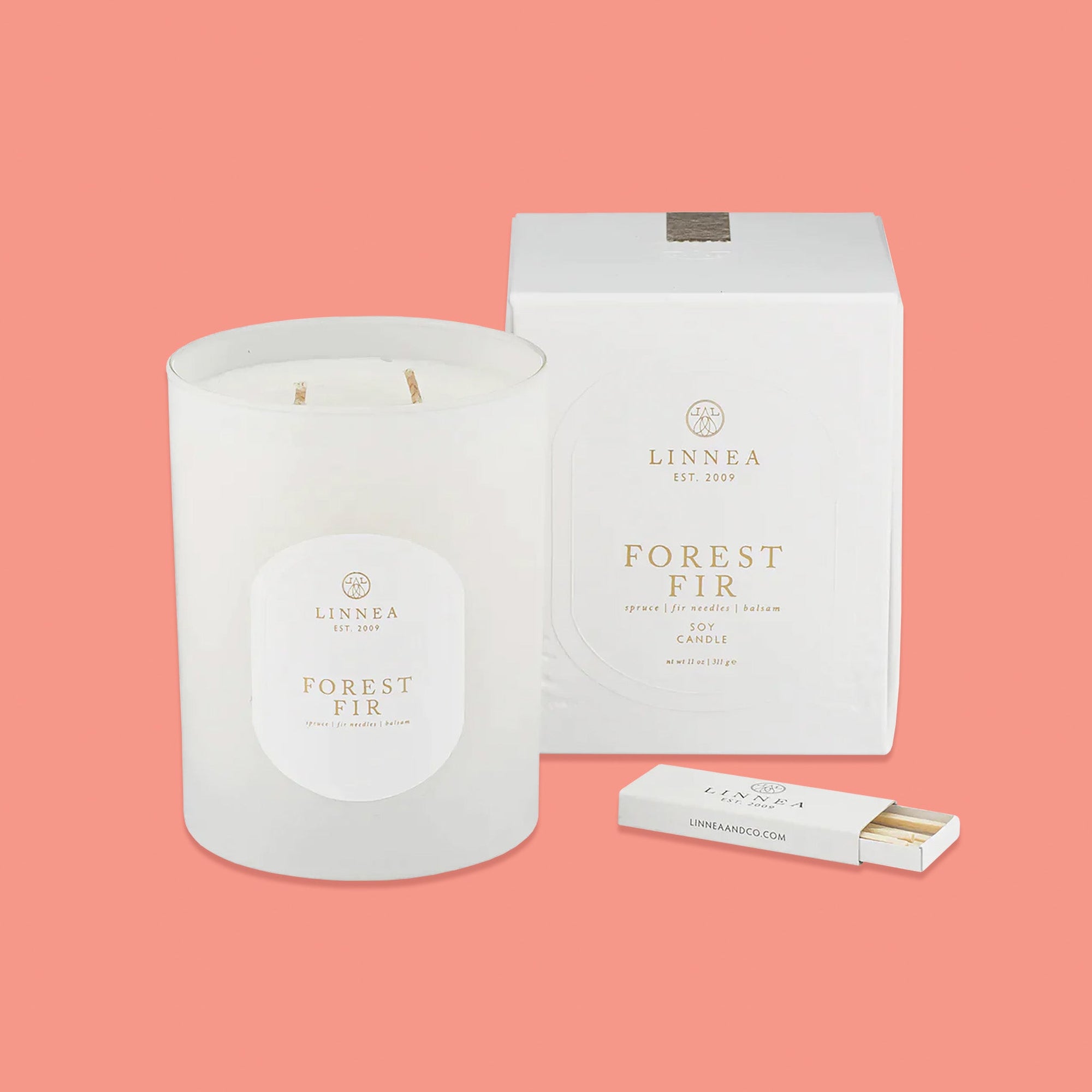 On a pink coral background sits a frosted glass candle with a white box next to it. In front of the box is a white box of matches. It says in gold lettering "LINNEA EST 2009", "FOREST FIR spruce | fir needles | balsam SOY CANDLE." nt wt 11 oz | 311 g