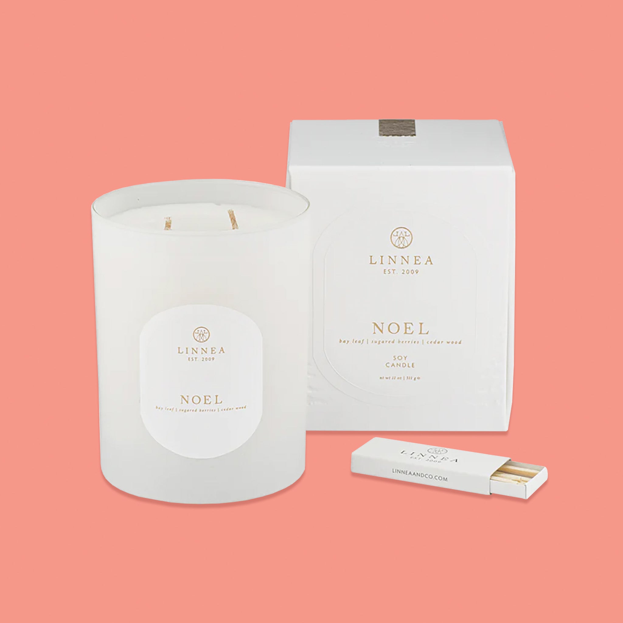 On a pink coral background sits a frosted glass candle with a white box next to it. In front of the box is a white box of matches. It says in gold lettering "LINNEA EST 2009", "NOEL bay leaf | sugar berries | cedar wood SOY CANDLE." nt wt 11 oz | 311 g