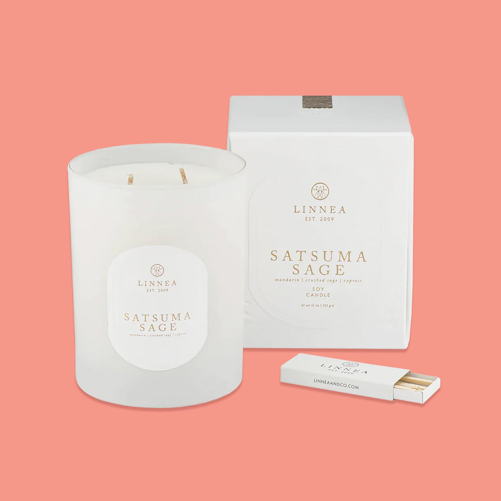 On a pink coral background sits a frosted glass candle with a white box next to it. In front of the box is a white box of matches. It says in gold lettering "LINNEA EST 2009", "SATSUMA SAGE mandarin | crushed sage | cypress SOY CANDLE." nt wt 11 oz | 311 g