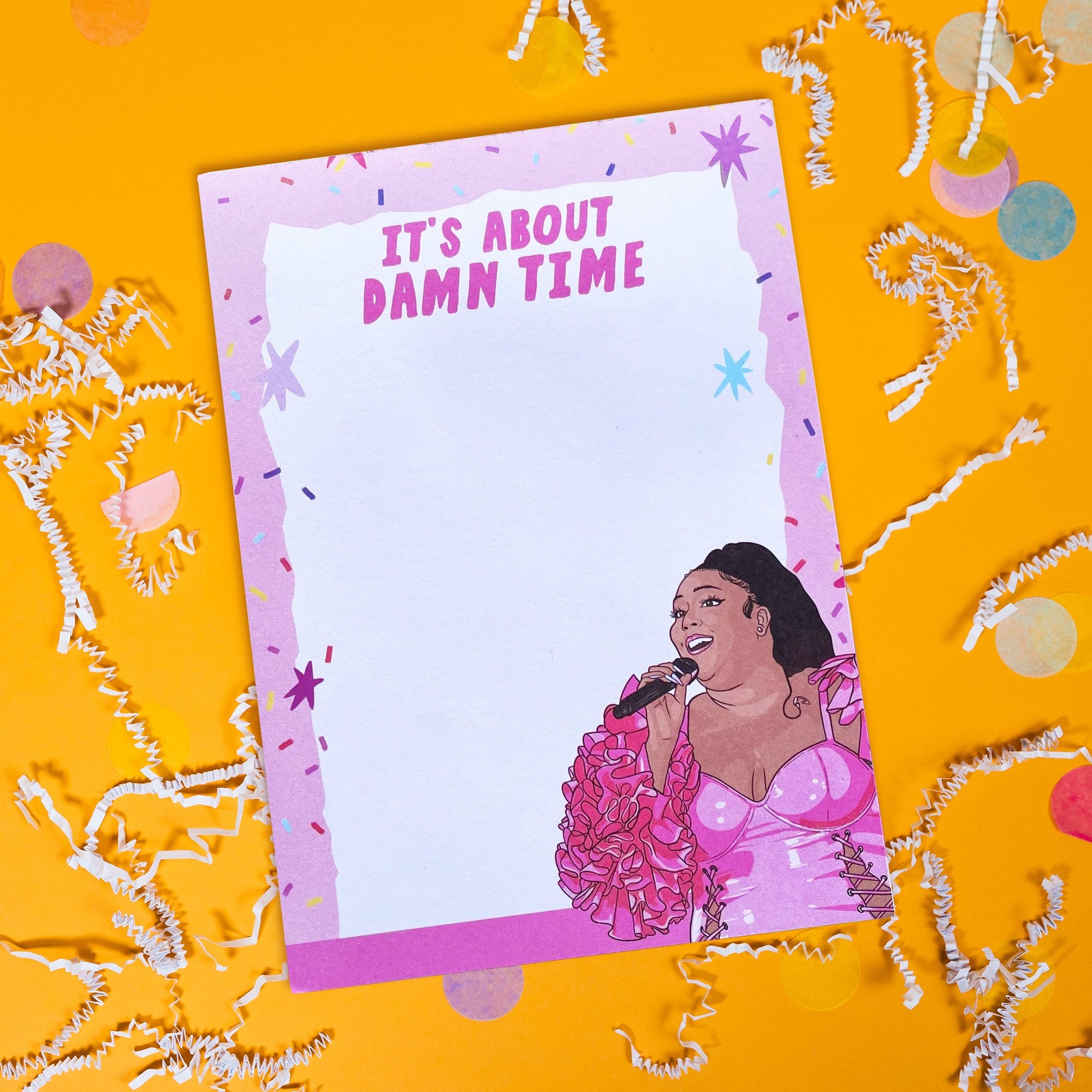 On a sunny mustard background sits a notepad with white crinkle and big, colorful confetti scattered around.. This Lizzo inspired notepard is colorful with different shades of pinks. There is a light pink border with colorful confetti and stars and a white, handdrawn box on top. At the top it says "IT'S ABOUT DAMN TIME" in hot pink, handwritten lettering. At the bottom right corner is an illustration of Lizzo with a hot pink outfit on and a microphone in her hand.