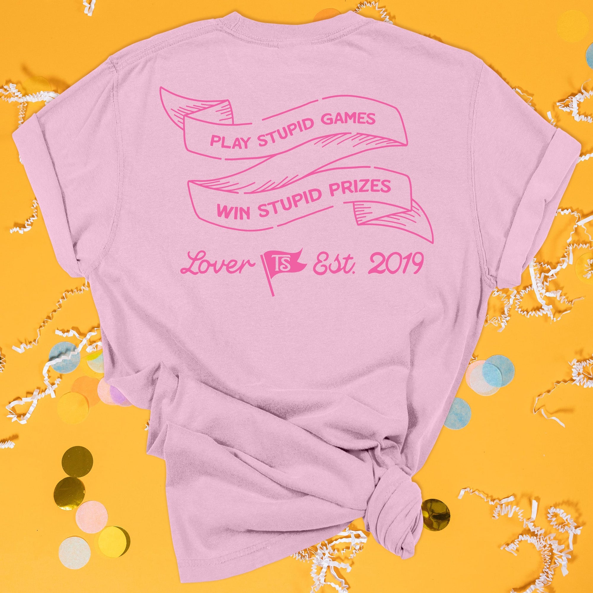 On a sunny mustard background sits the back of a t-shirt with white crinkle and big, colorful confetti scattered around. This Taylor Swift Inspired Reputation tee is pink with bubblegum lettering and ribbon illustration. In the ribbon it says "PLAY STUPID GAMES WIN STUPID PRIZES" in all caps. Under it says "Lover Est. 2019" in script lettering. There is a flag that has "TS" in it. 
