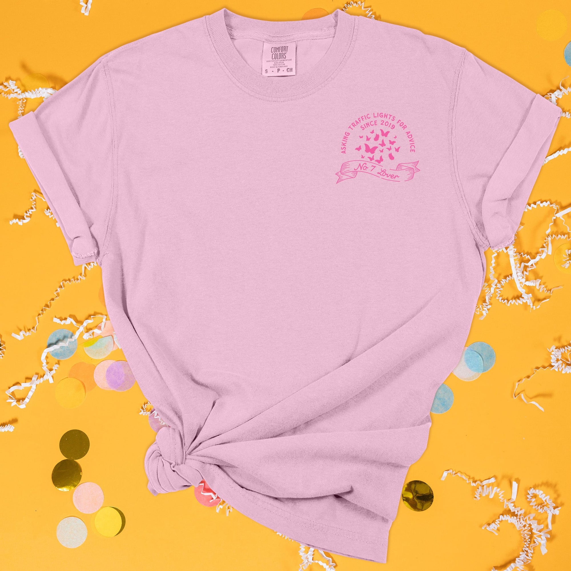 On a sunny mustard background sits the front of a t-shirt with white crinkle and big, colorful confetti scattered around. This Taylor Swift Inspired Reputation tee is pink with bubblegum lettering and illustrations. There are butterflies with a ribbon under it over the left chest area. It says "ASKING TRAFFIC LIGHTS FOR ADVICE SINCE 2019" in all caps and "No. 7 Lover" in script lettering.