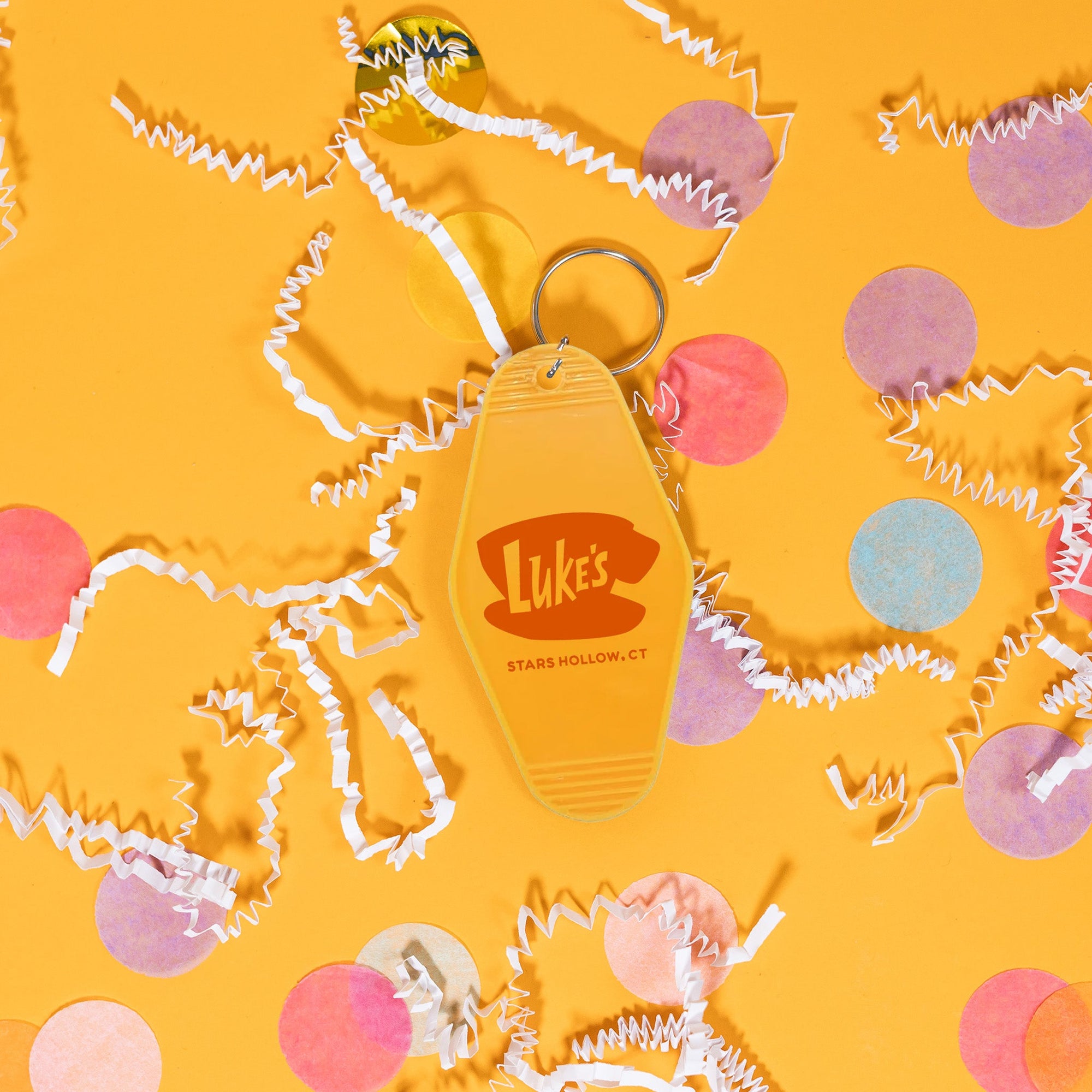 On a sunny mustard background sits a key chain with white crinkle and big, colorful confetti scattered around. This Gilmore Girls inspired mustard yellow vintage hotel key chain has a red illustration of Luke's Stars Hollow.