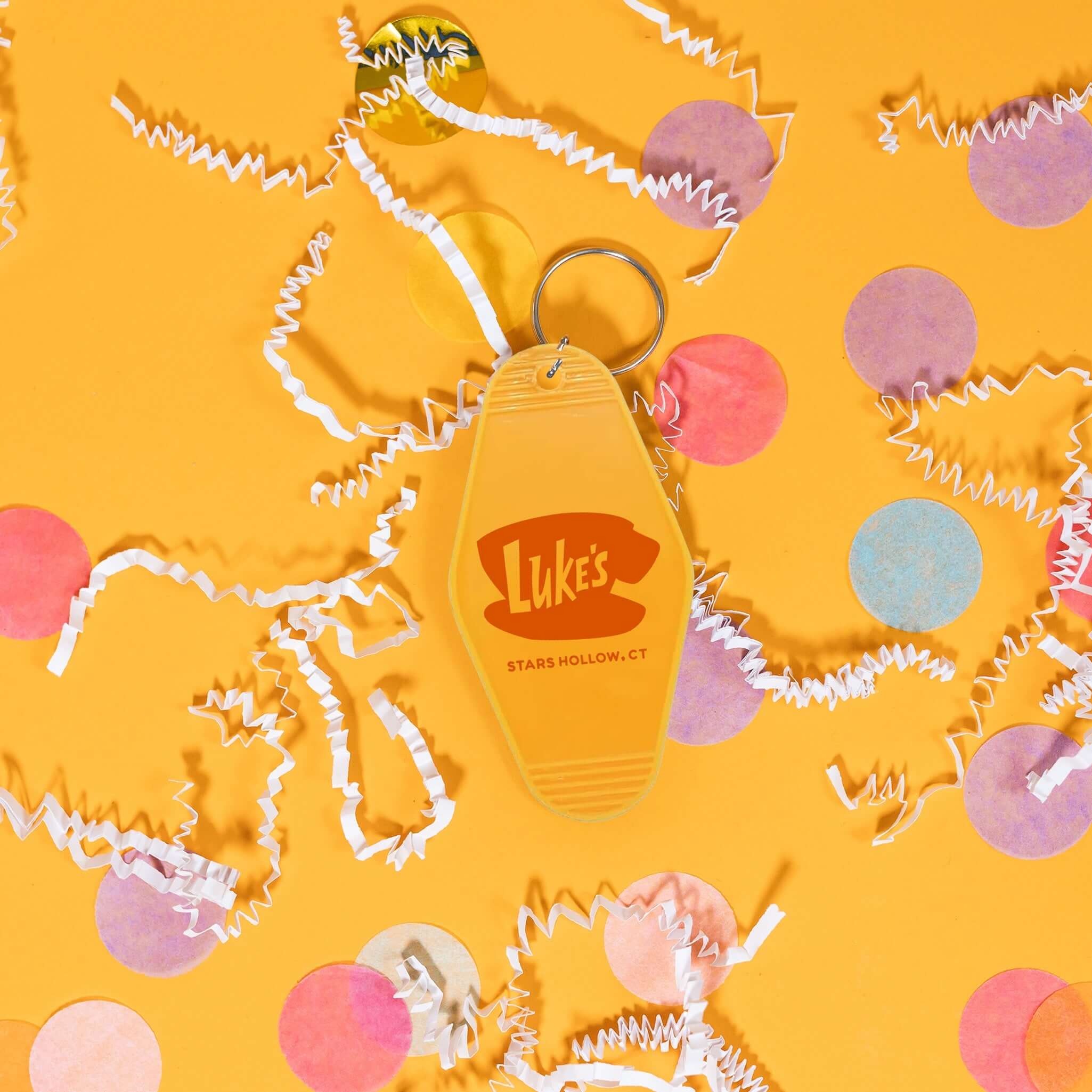 On a sunny mustard background sits a key chain with white crinkle and big, colorful confetti scattered around. This Gilmore Girls inspired mustard yellow vintage hotel key chain has a red illustration of Luke's Stars Hollow.