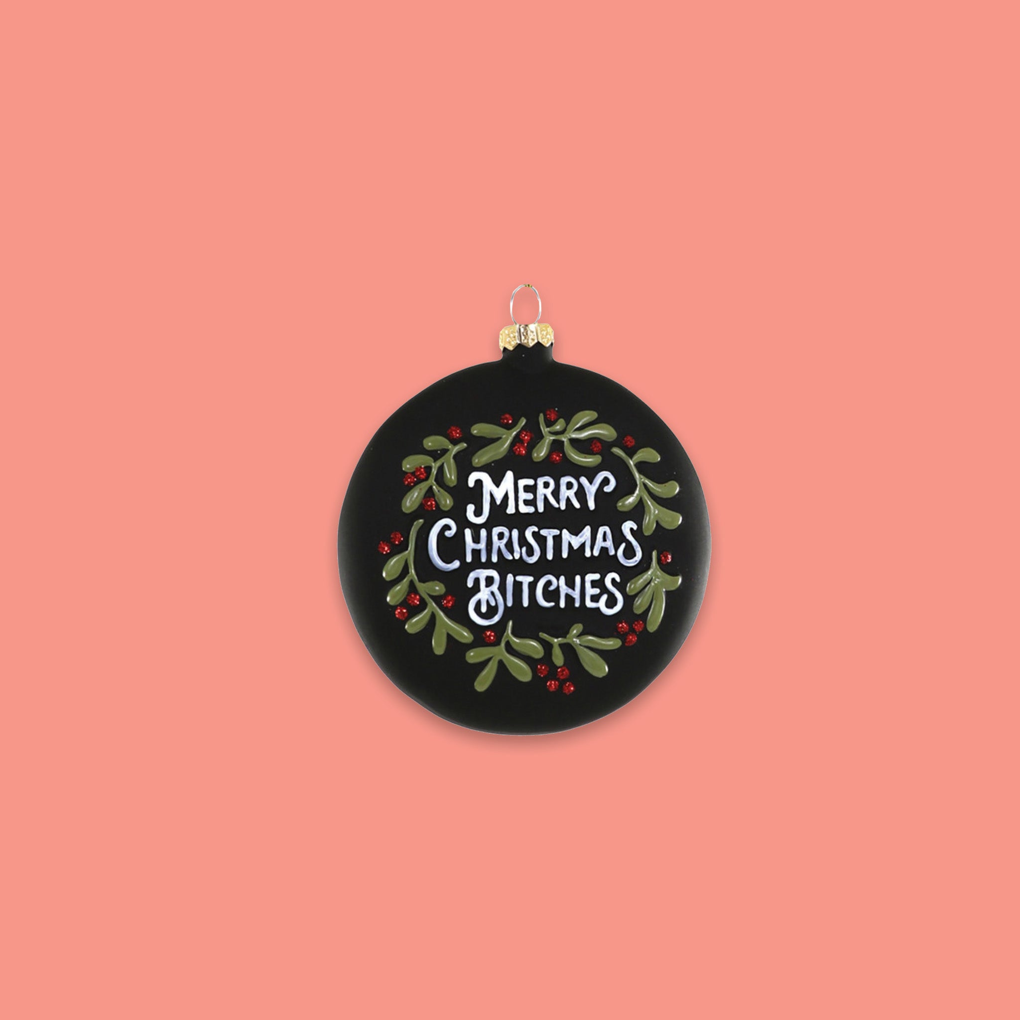 On a coral pink background sits a round ornament. This glass ornament is black with green and red hollies and berries and it says "MERRY CHRISTMAS BITCHES" in white lettering.