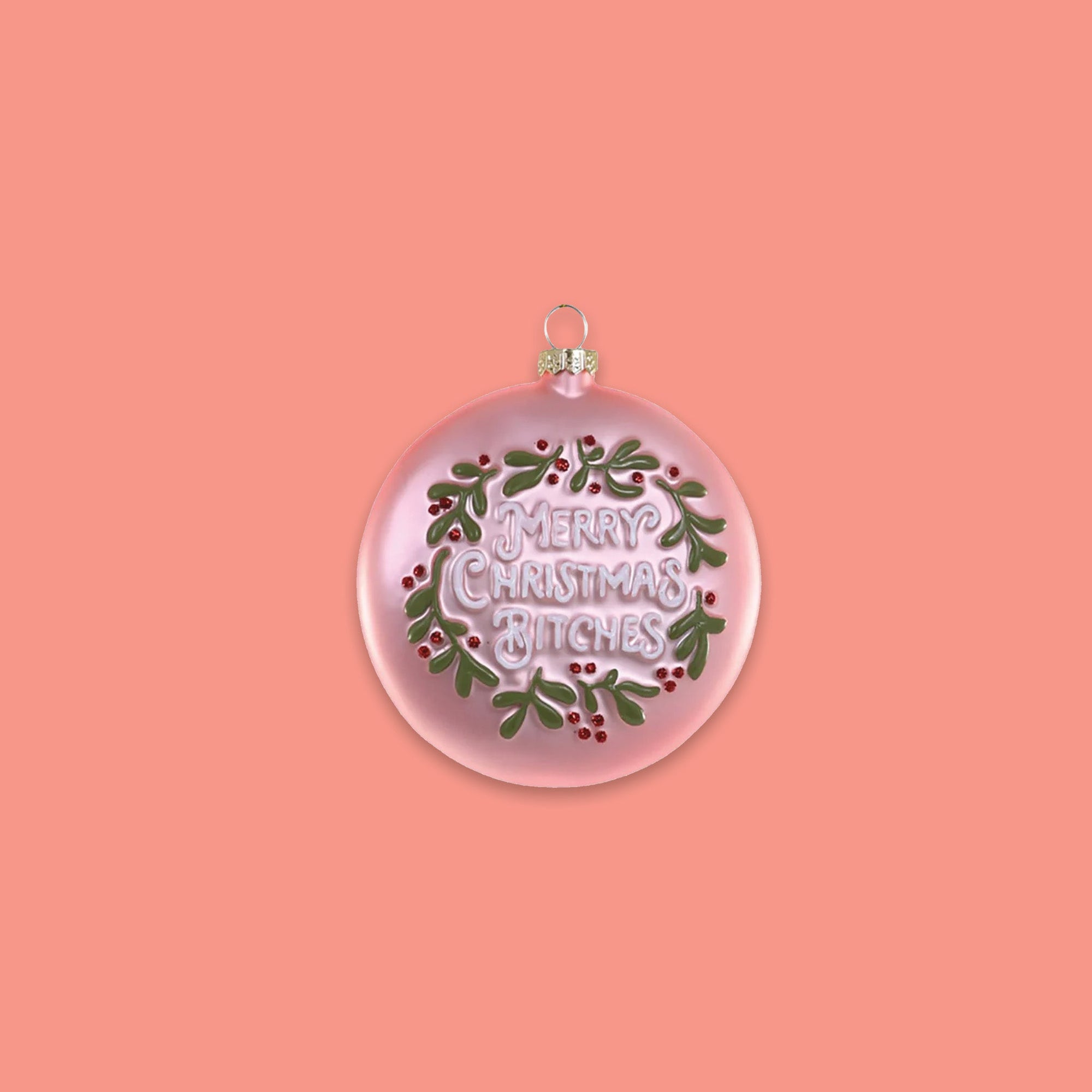 On a coral pink background sits a round ornament. This glass ornament is pink with green and red hollies and berries and it says "MERRY CHRISTMAS BITCHES" in white lettering.