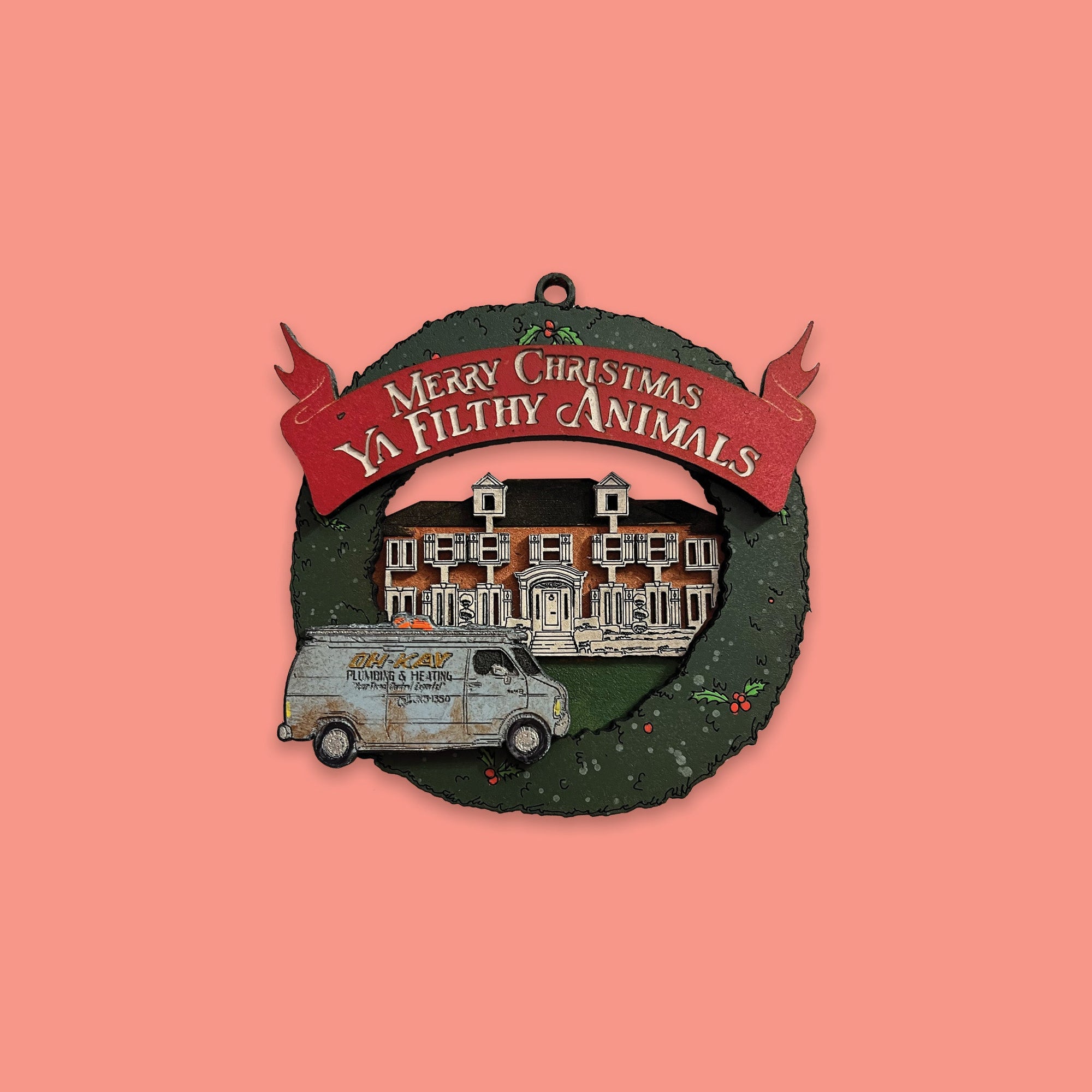 On a coral pink background sits a wreath ornament. This Home Alone inspired wood ornament is a green wreath with Kevin McAllister's house and the blue OH-KAY PLUMBING AND HEATING in front ot it. At the top is a red banner that says "MERRY CHRISTMAS YA FILTHY ANIMALS" in white lettering.