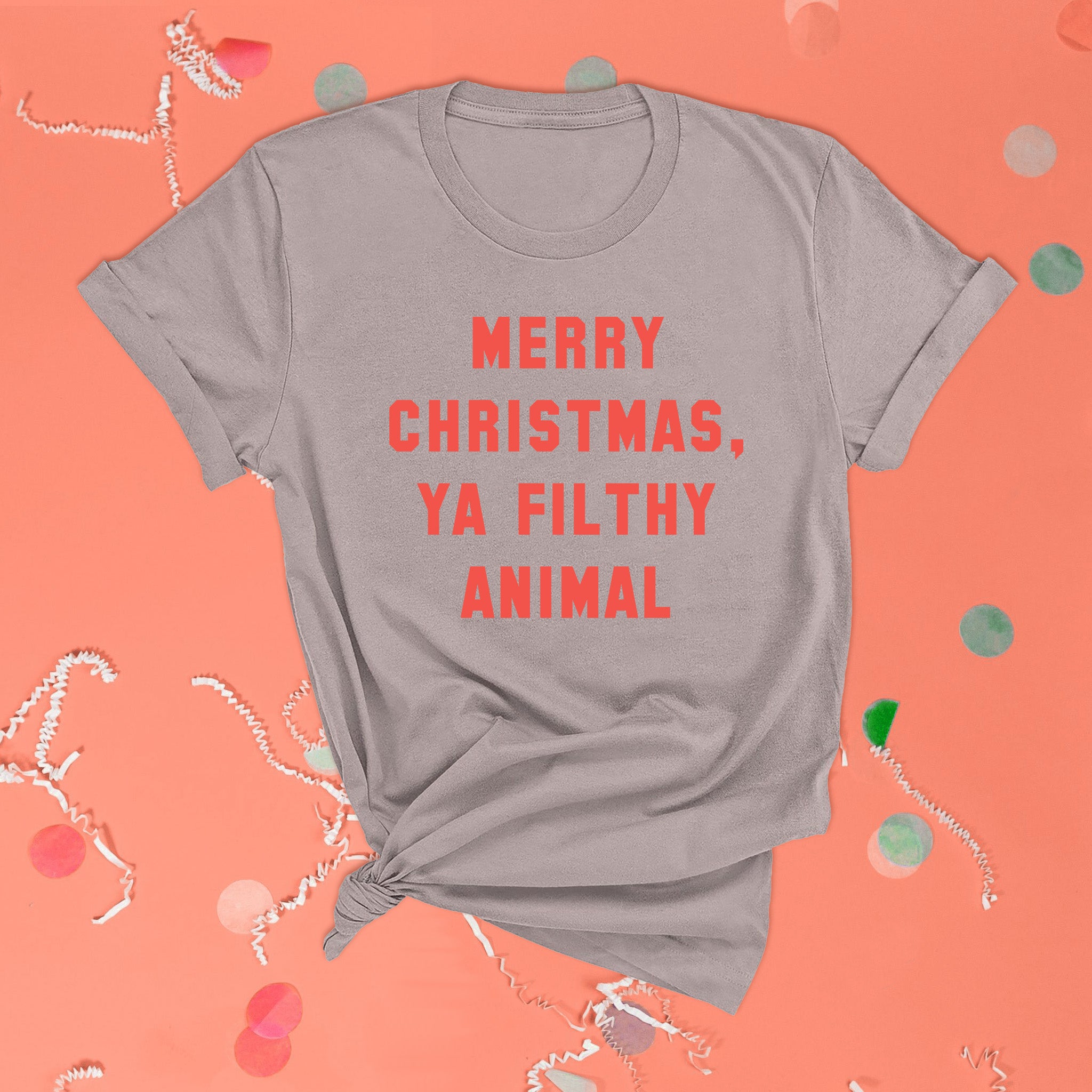 On a pink coral background sits the front of a t-shirt with white crinkle and big, colorful confetti scattered around. This Home Alone Inspired crewneck grey t-shirt has bright coral red collegiate lettering and it says "MERRY CHRISTMAS, YA FILTHY ANIMAL."