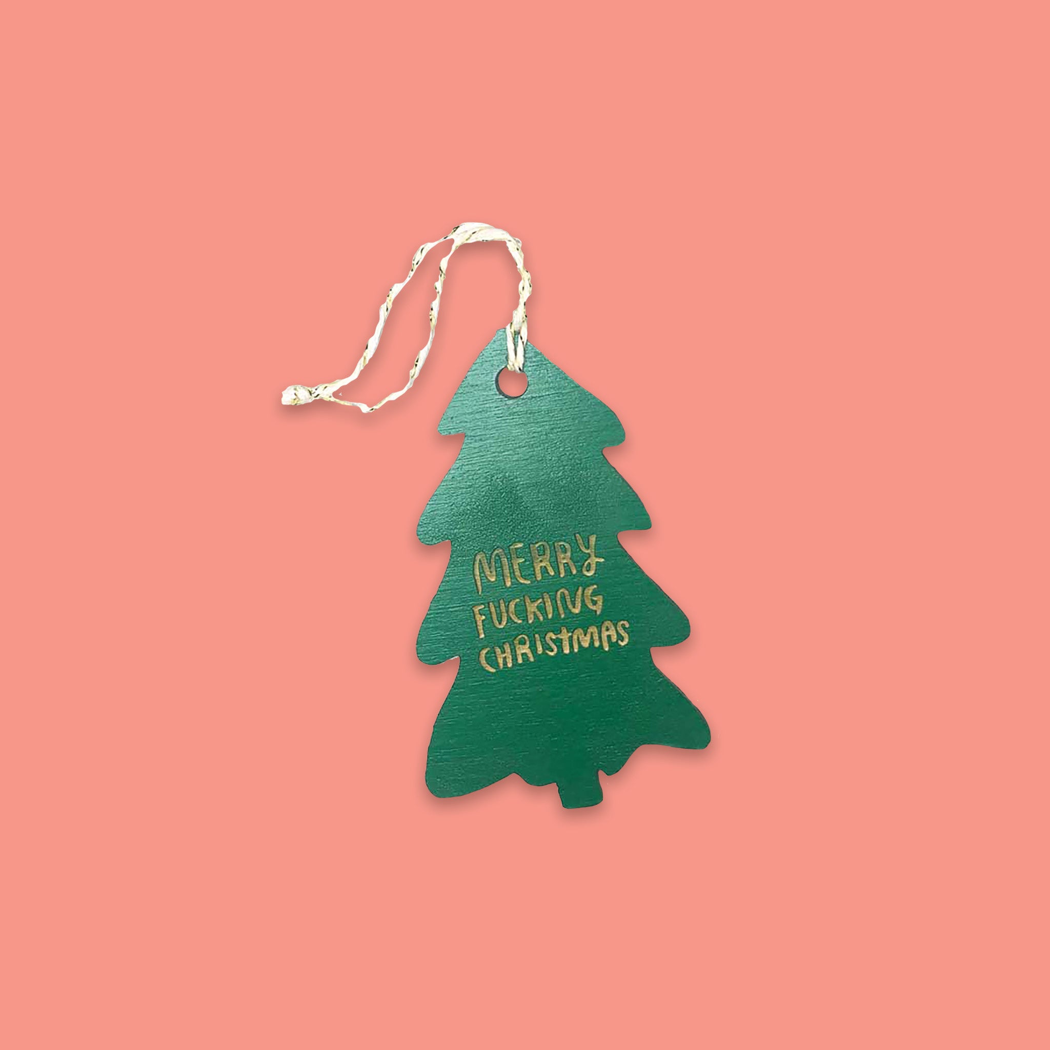 On a coral pink background sits a tree ornament. This wood ornament is in the shap of a christmas tree and it says in gold lettering "MERRY FUCKING CHRISTMAS."