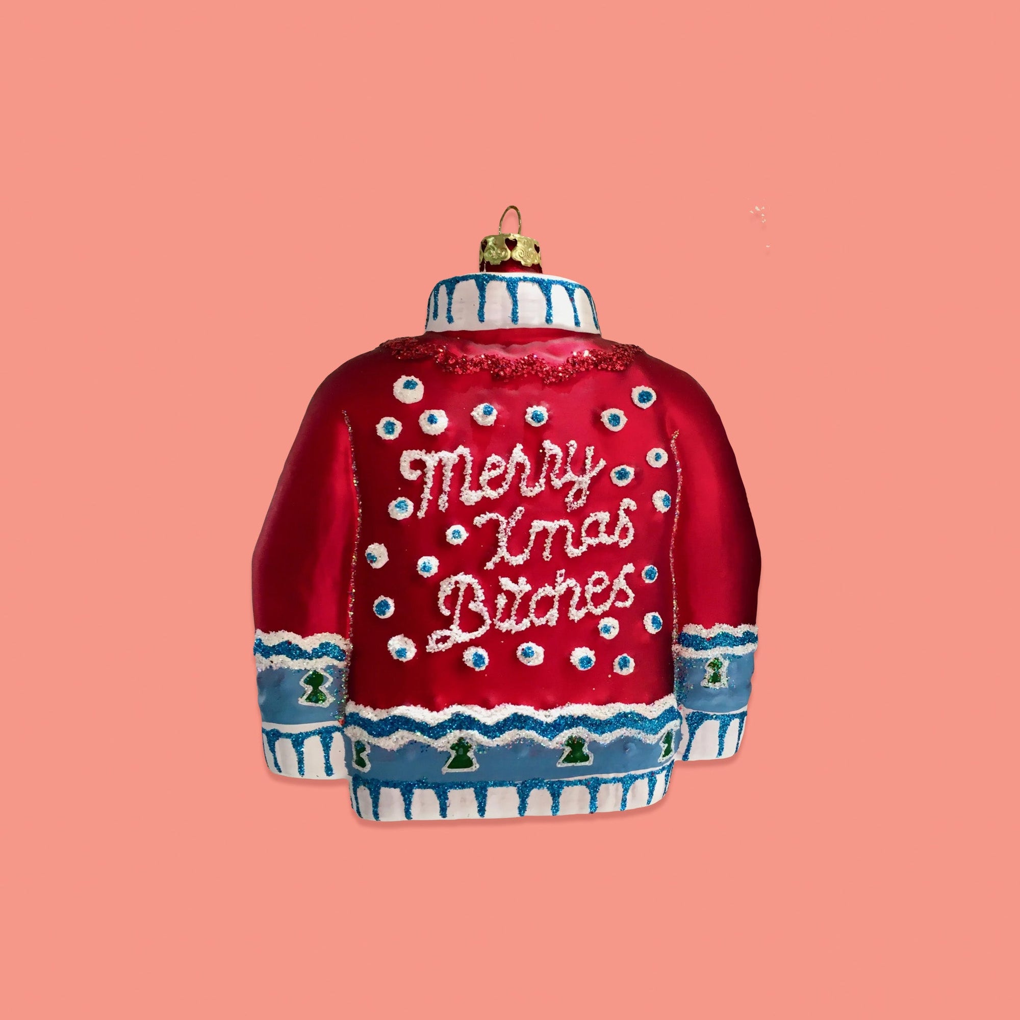 On a coral pink background sits an ornament. This ugly sweater inspired glass ornament is red and blue with white dpts and it says "Merry Xmas Bitches" in white glitter script lettering. There is red, white and blue glitter.