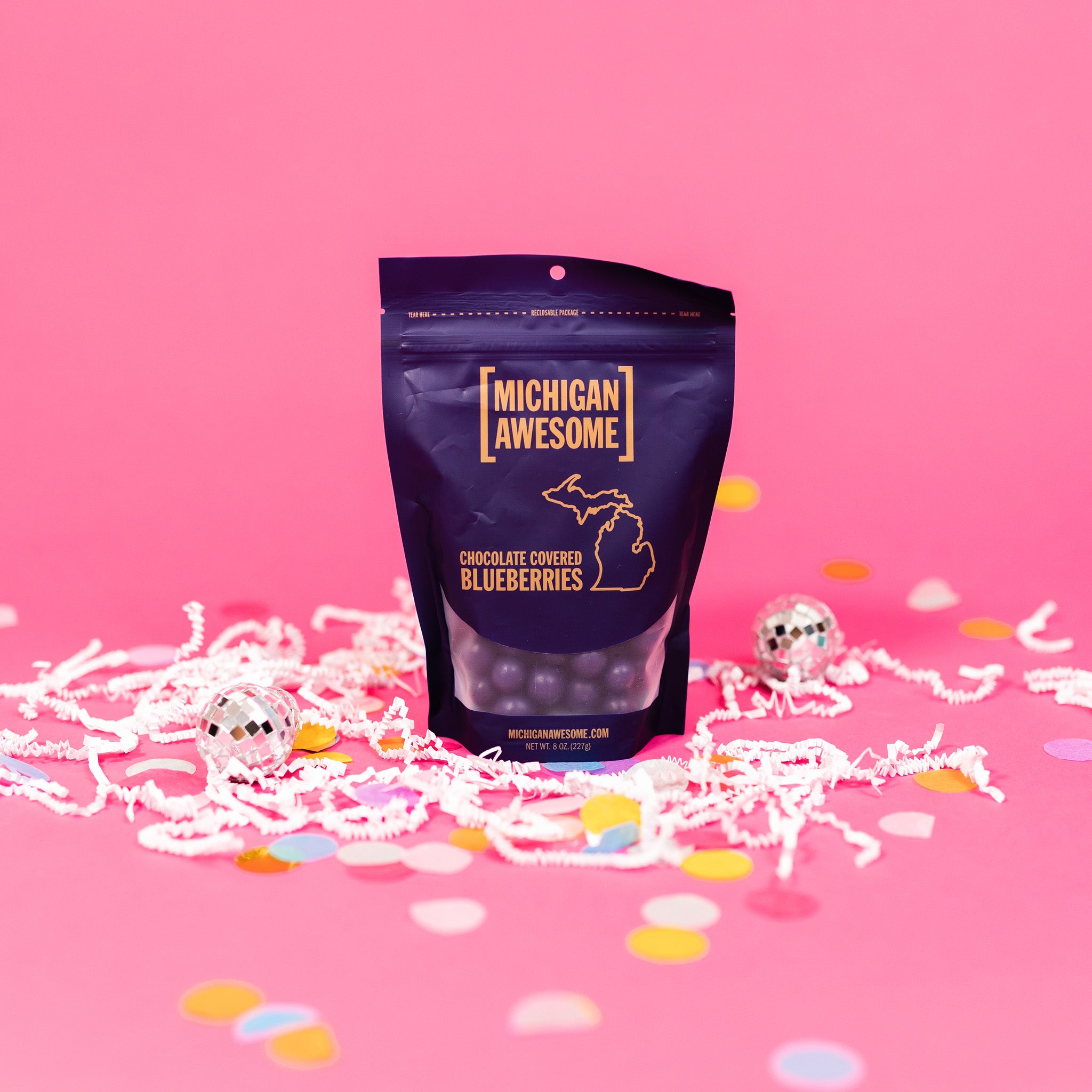 On a bubblegum pink background sits a bag with white crinkle and big, colorful confetti scattered around. There are mini disco balls. It is filled with chocolate covered blueberries. It says "MICHIGAN AWESOME," and "CHOCOLATE COVERED BLUEBERRIES" in goldish-yellow, all caps block font. It has an outlined illustration of the state of Michigan-UP. NET WT. 8 OZ. (227G)