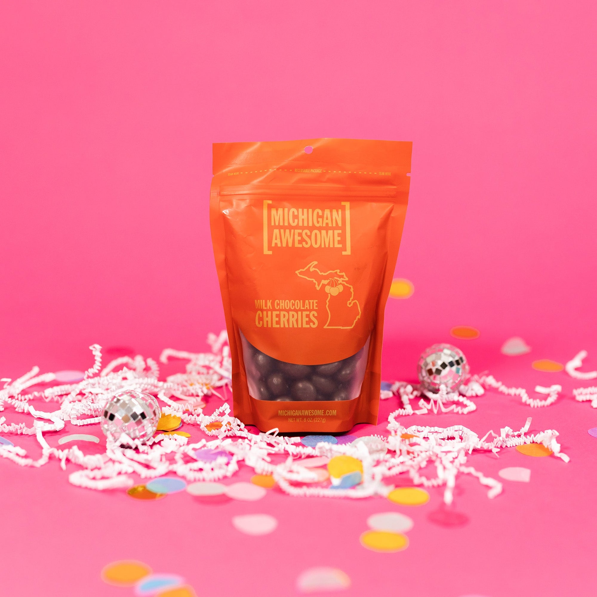 On a bubblegum pink background sits a bag with white crinkle and big, colorful confetti scattered around. There are mini disco balls. This red package has chocolate covered cherries in the package. It says in gold "MICHIGAN AWESOME," "MILK CHOCOLATE CHERRIES" in all caps block font. It also says "MICHIGANAWESOME.COM" in gold, all caps block font. NEW WT. 8 OZ. (227g)