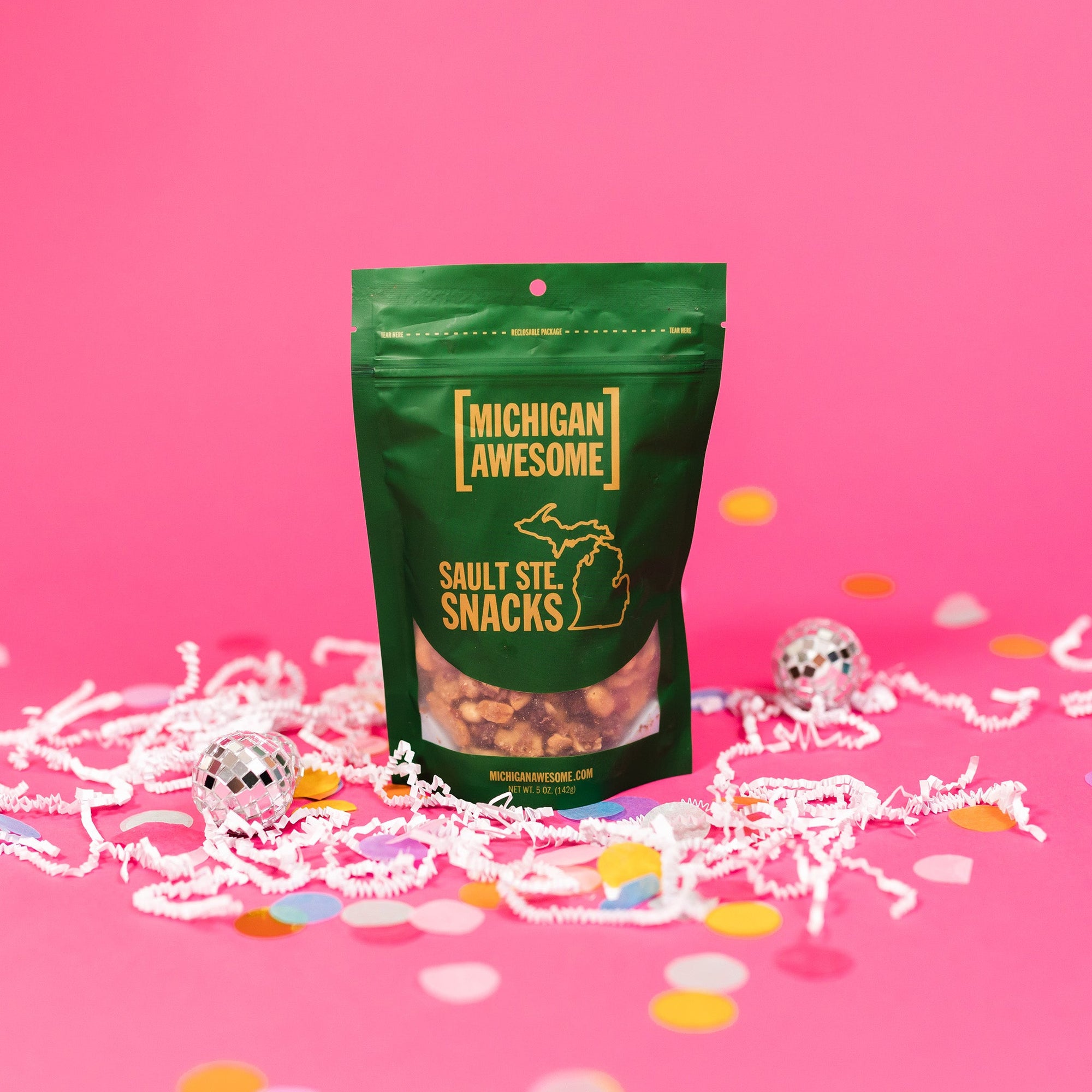 On a bubblegum pink background sits a bag with white crinkle and big, colorful confetti scattered around. There are mini disco balls. This green bag filled with sesame sticks, sesame seeds, and salt and pepper peanuts. It says "MICHIGAN AWESOME," and "SAULT STE. SNACKS" in goldish-yellow, all caps block font. It has an outlined illustration of the state of Michigan-UP. 