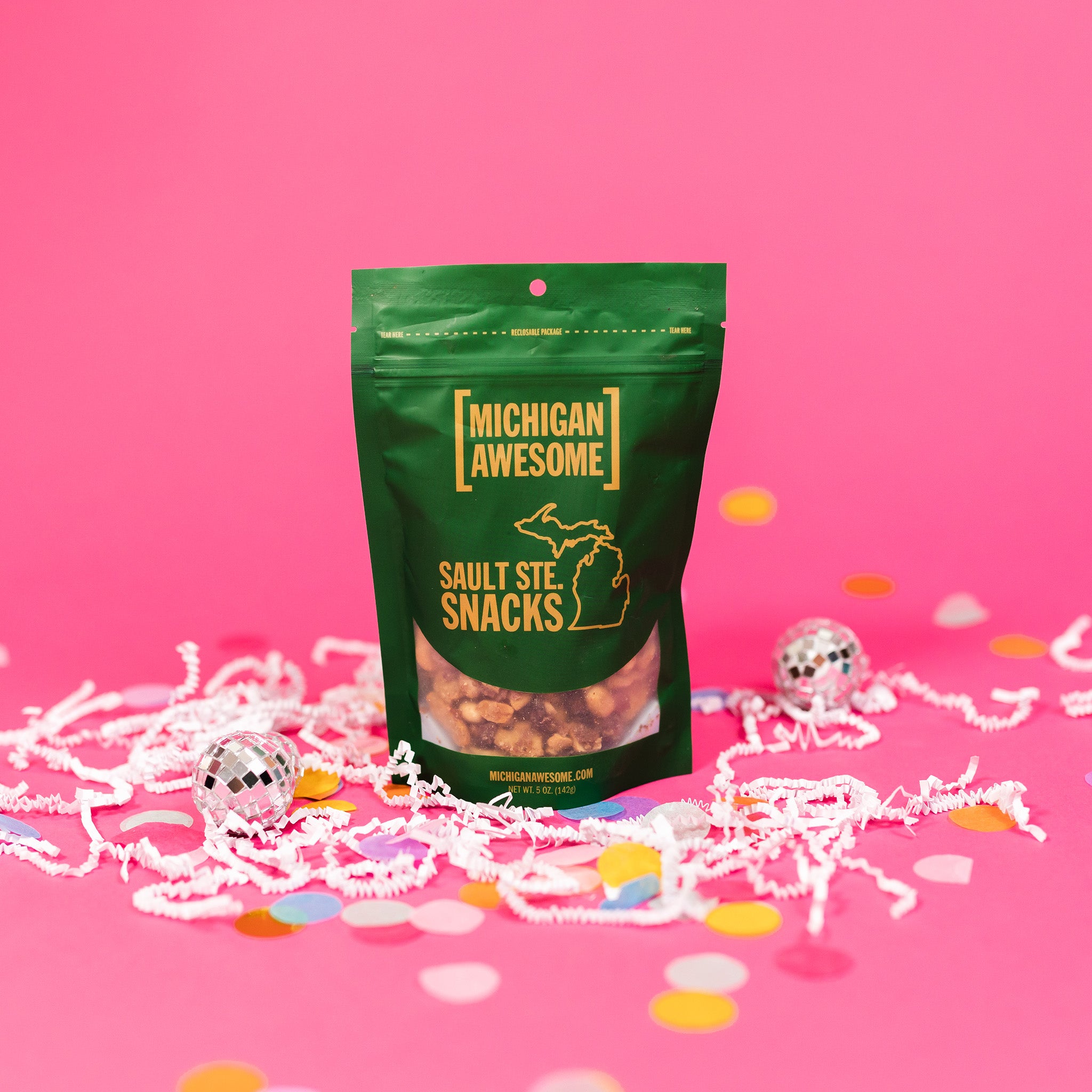 On a bubblegum pink background sits a bag with white crinkle and big, colorful confetti scattered around. There are mini disco balls. This green bag filled with sesame sticks, sesame seeds, and salt and pepper peanuts. It says "MICHIGAN AWESOME," and "SAULT STE. SNACKS" in goldish-yellow, all caps block font. It has an outlined illustration of the state of Michigan-UP. 