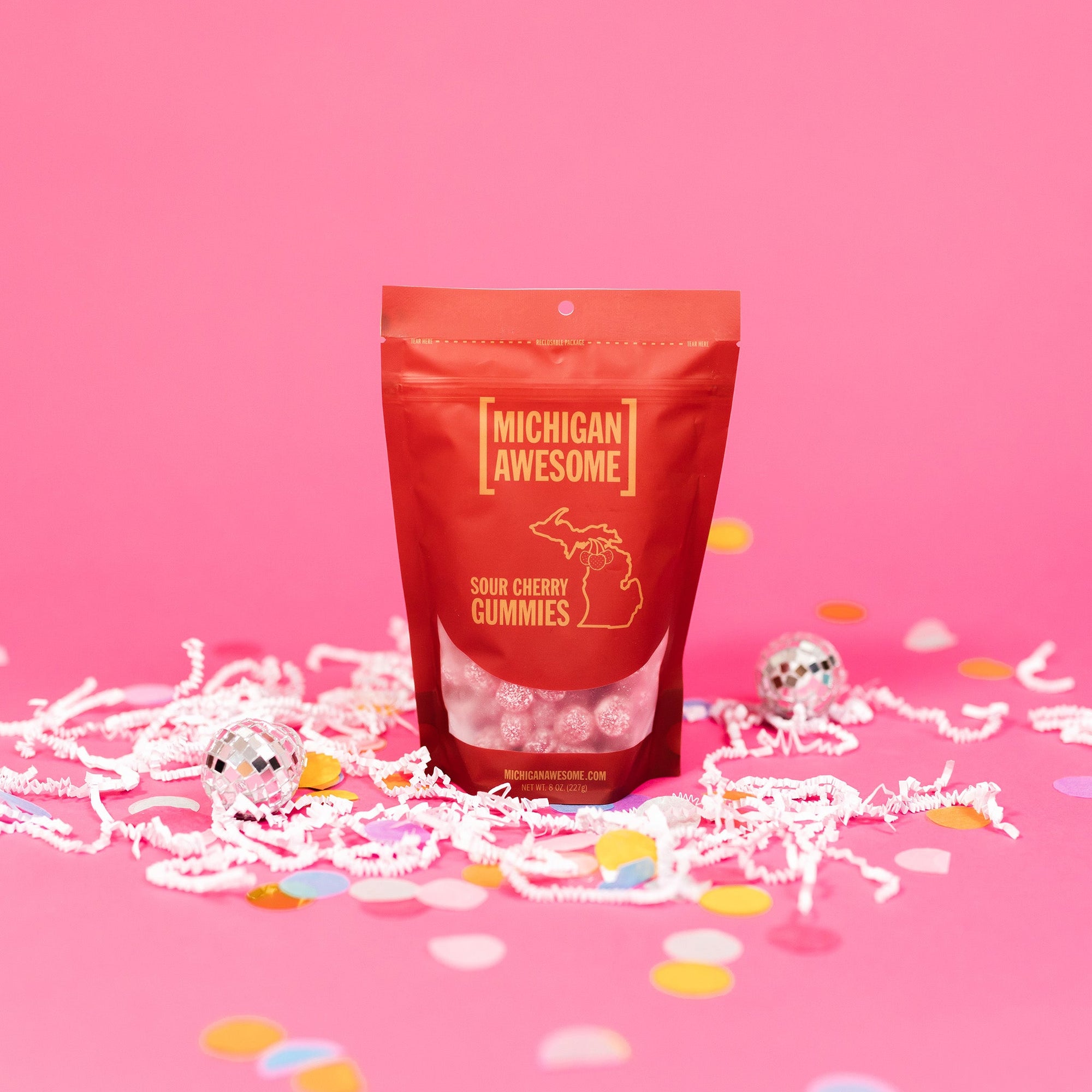 On a bubblegum pink background sits a bag with white crinkle and big, colorful confetti scattered around. There are mini disco balls. This red bag filled with sugar sour cherry gummies. It says "MICHIGAN AWESOME," and "SOUR CHERRY GUMMIES" in goldish-yellow, all caps block font. It has an outlined illustration of the state of Michigan-UP with cherries. There are sugar sour cherry gummies sitting around the bag.