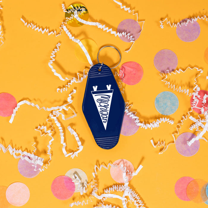On a sunny mustard background sits a keychain with white crinkle and big, colorful confetti scattered around. This navy, vintage motel keychain has a white pennant on it and it says "Michigan" in navy, script handwritten lettering.