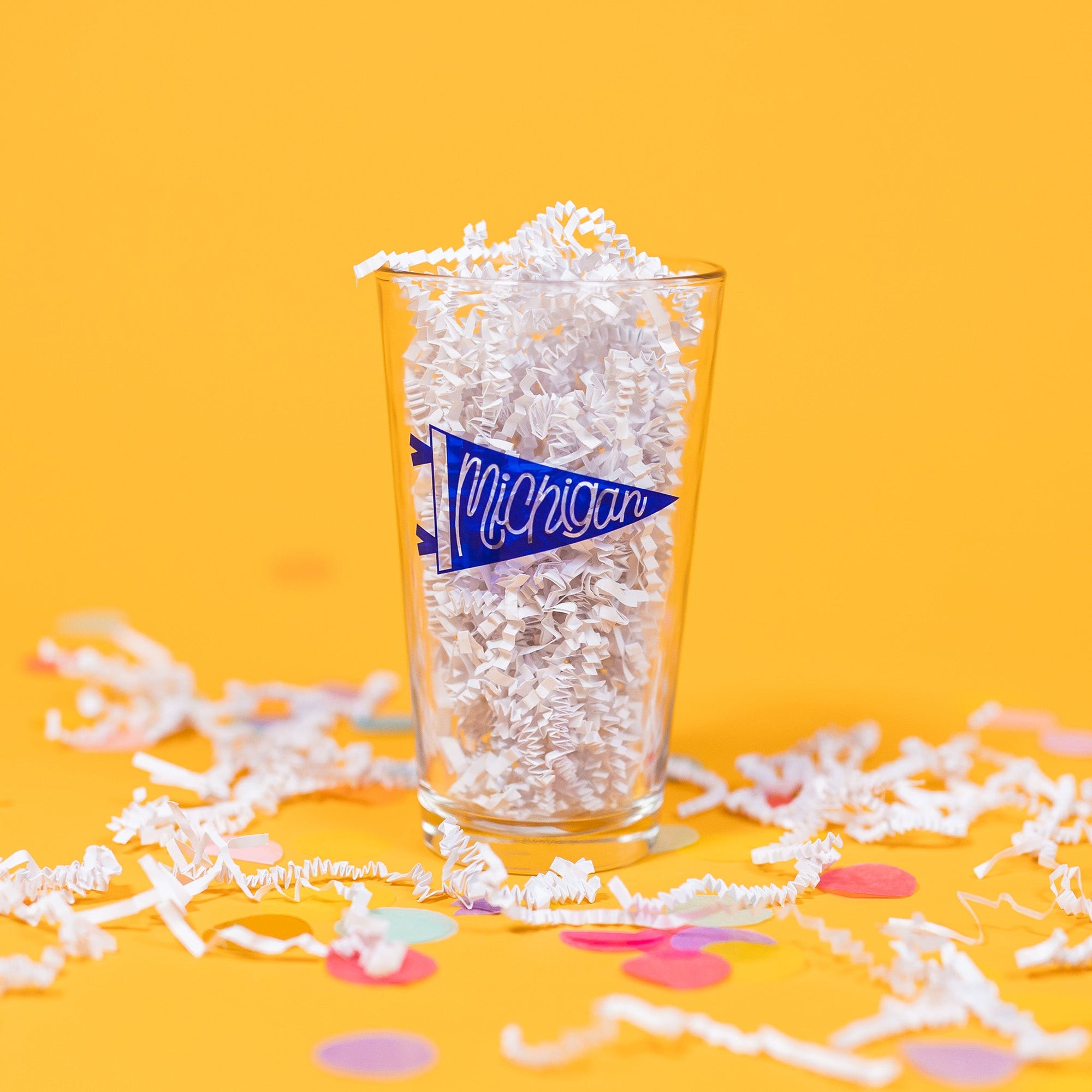 On a sunny mustard background sits a pint glass with white crinkle and big, colorful confetti scattered around. This beer pint glass is filled with white crinkle and it has a blue pennant that says "Michigan" in script hand lettering.