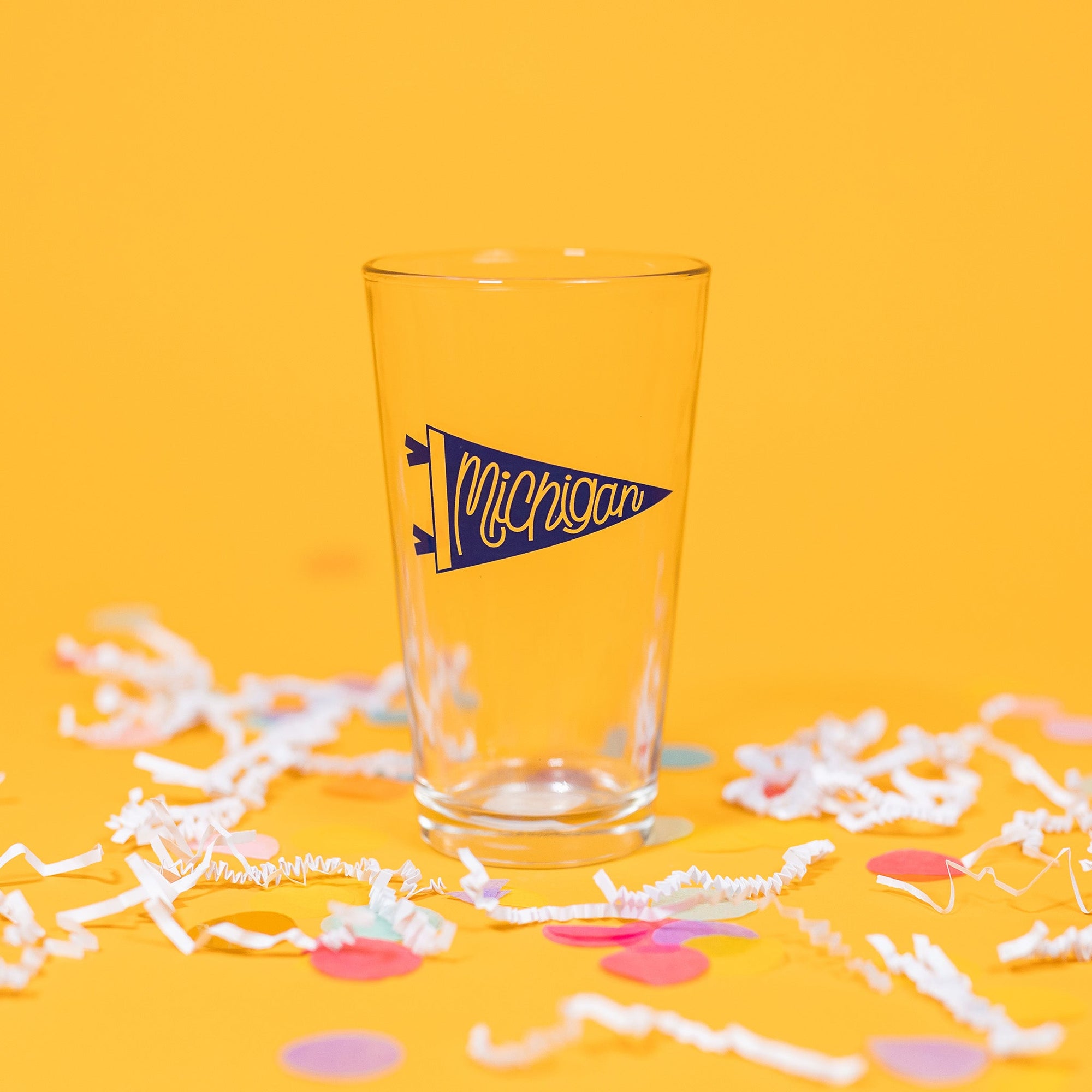 On a sunny mustard background sits a pint glass with white crinkle and big, colorful confetti scattered around. This beer pint glass has a blue pennant that says "Michigan" in script hand lettering.