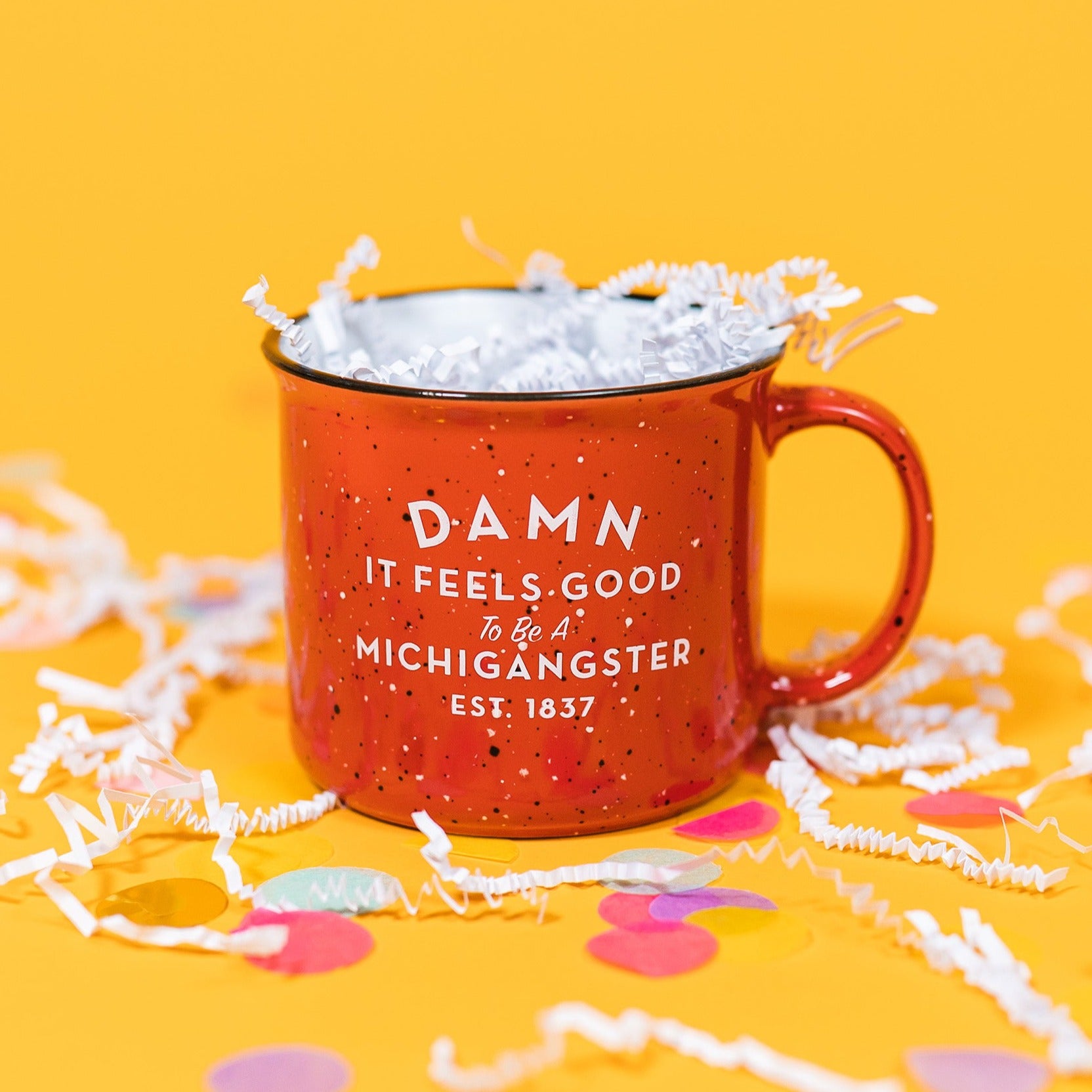 On a sunny mustard background sits a mug with white crinkle and big, colorful confetti scattered around. The red campfire mug has black and white flecks with a black rim and the inside is white. It says "DAMN IT FEELS GOOD To Be A MICHIGANGSTER" and "EST. 1837" in a white, block font. It is filled with white crinkle.