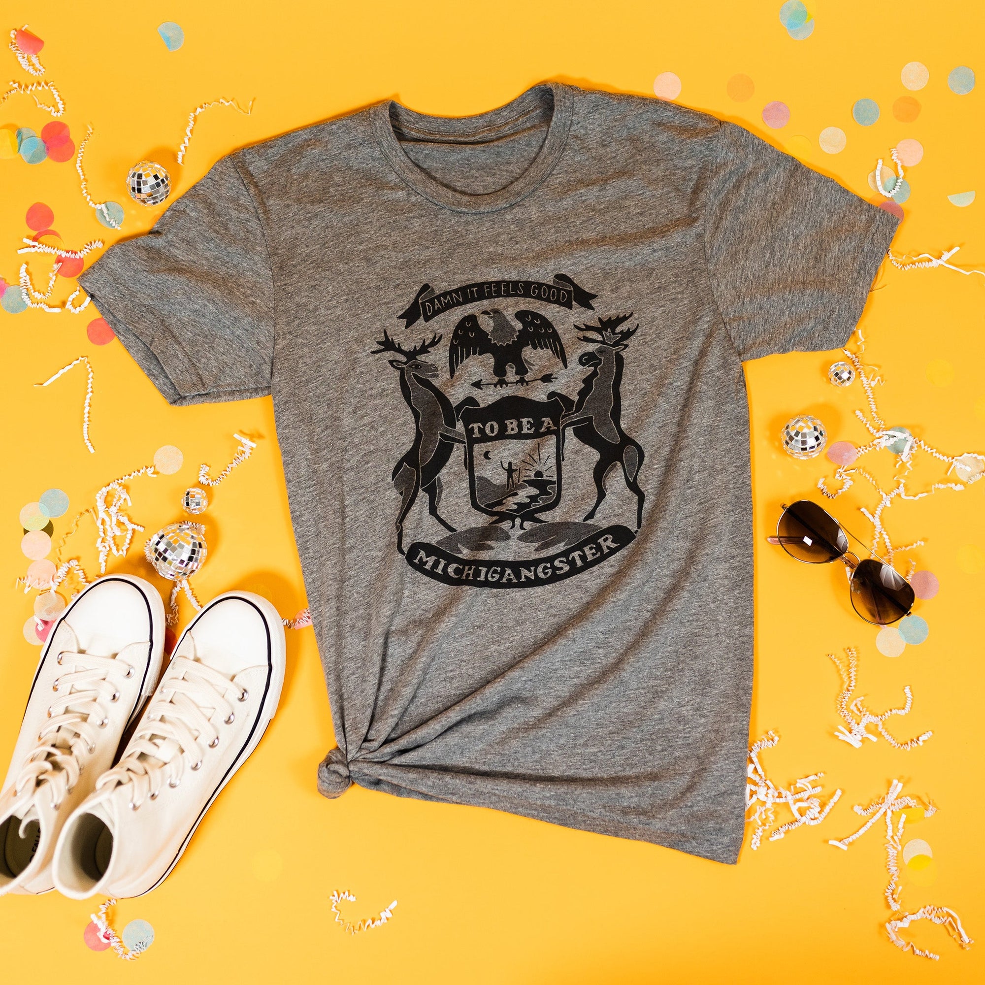 On a sunny mustard background sits a t-shirt with white crinkle and big, colorful confetti scattered around. There are mini disco balls, sunglasses, and a pair of white sneakers. This heather grey tee features hand lettering and custom illustrations of a modern, lighthearted take on the flag of Michigan. It is in black ink.