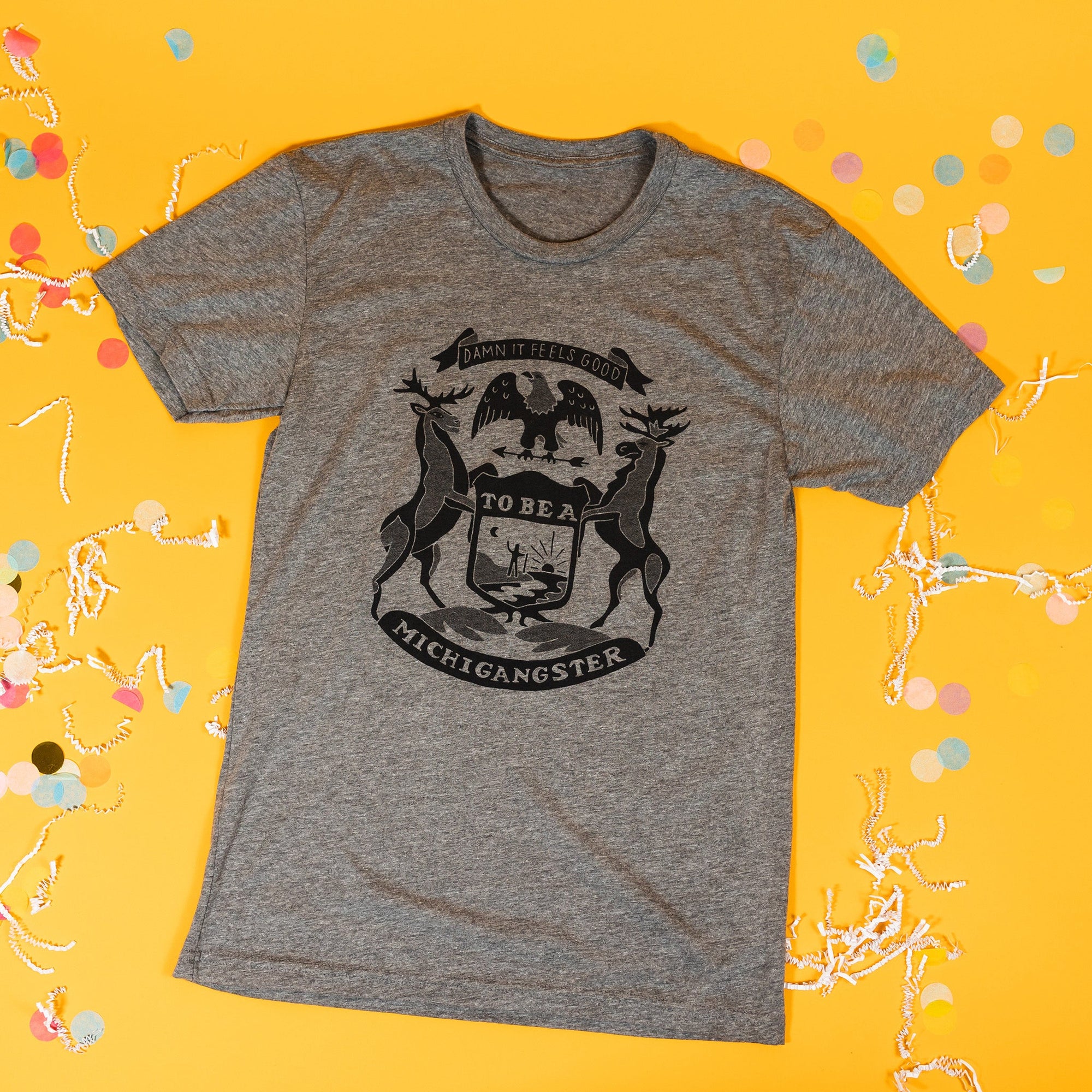 On a sunny mustard background sits a t-shirt with white crinkle and big, colorful confetti scattered around. This heather grey tee features hand lettering and custom illustrations of a modern, lighthearted take on the flag of Michigan. It is in black ink.