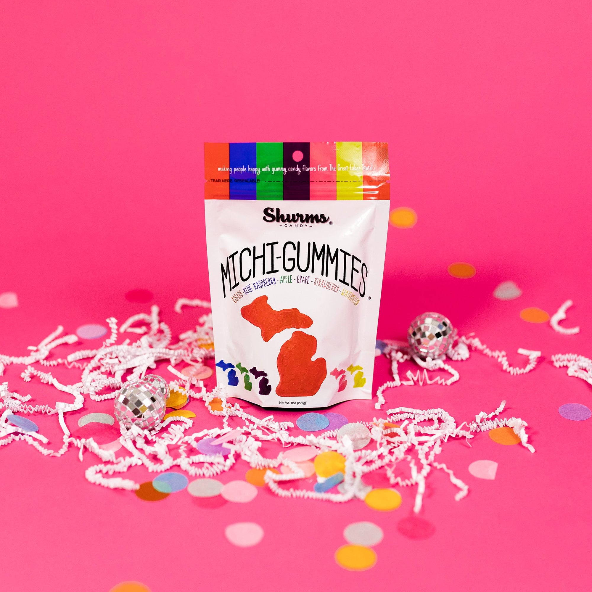 On a bubblegum pink background sits a bag with white crinkle and big, colorful confetti scattered around. There are mini disco balls. This package is colorful and says "making people happy with hummy candy flavors from The Great Lakes State!" in white, handwritten lettering. It also says "MICHI-GUMMIES," "CHERRY • BLUE RASPBERRY • APPLE • STRAWBERRY • WATERMELON" in all caps, various colors font. It has illustrations of colored gummies in the the shape of Michigan state. Net Wt. 8oz (227g)