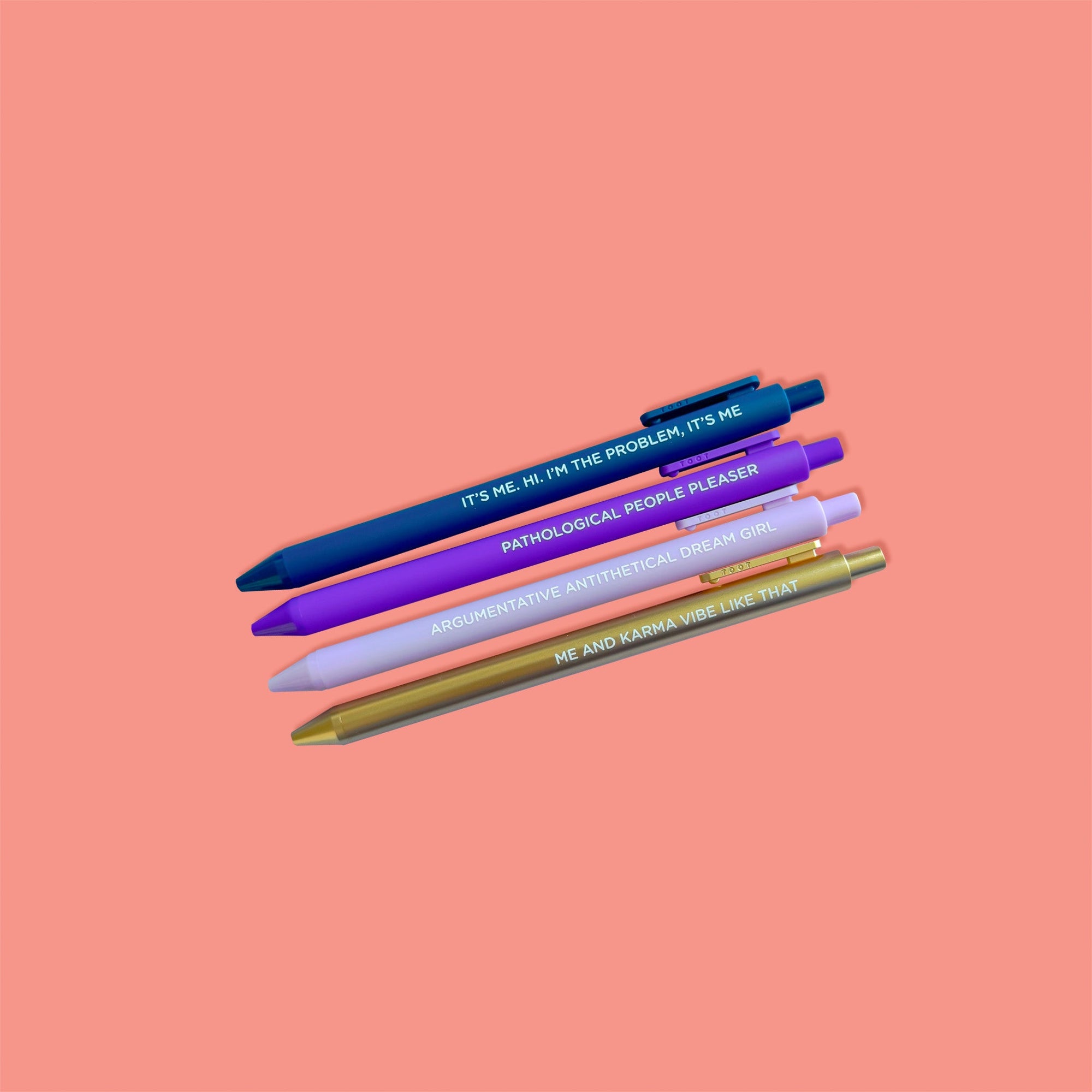 On a coral pink background sits a set of 4 pens. These Taylor Swift inspired pens are in colors of navy, purple, lavender, and gold. They say "IT'S ME, HI. I'M THE PROBLEM, IT'S ME", "PATHOLOGICAL PEOPLE PLEASER", " ARGUMENTATIVE ANTITHETICAL DREAM GIRL", "ME AND KARMA VIBE LIKE THAT."