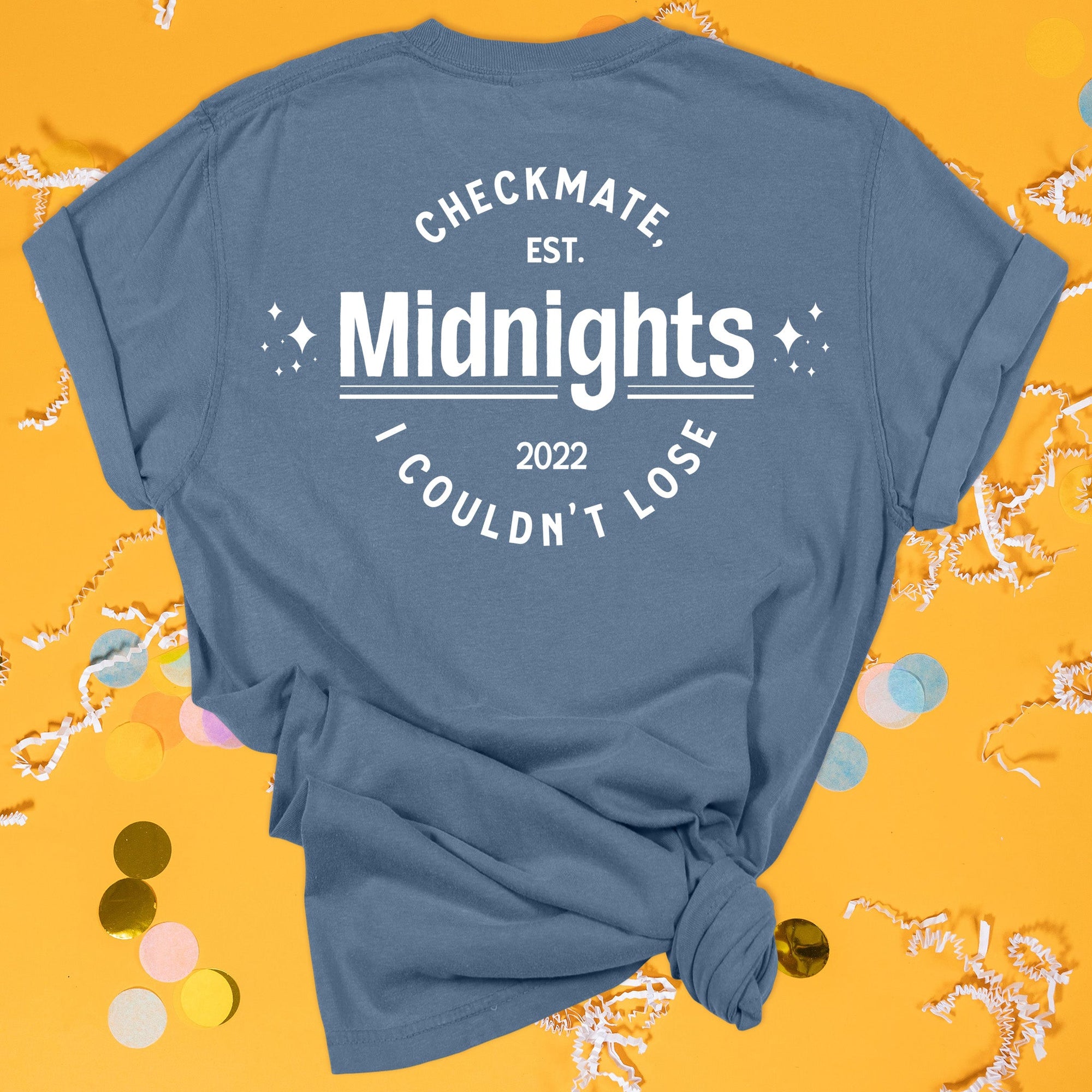 On a sunny mustard background sits the back of a t-shirt with white crinkle and big, colorful confetti scattered around. This Taylor Swift Inspired Reputation tee is dark blue with white lettering and sparkle stars. It says "CHECKMATE, I COULDN'T LOSE", "EST. 2022", and "Midnights."