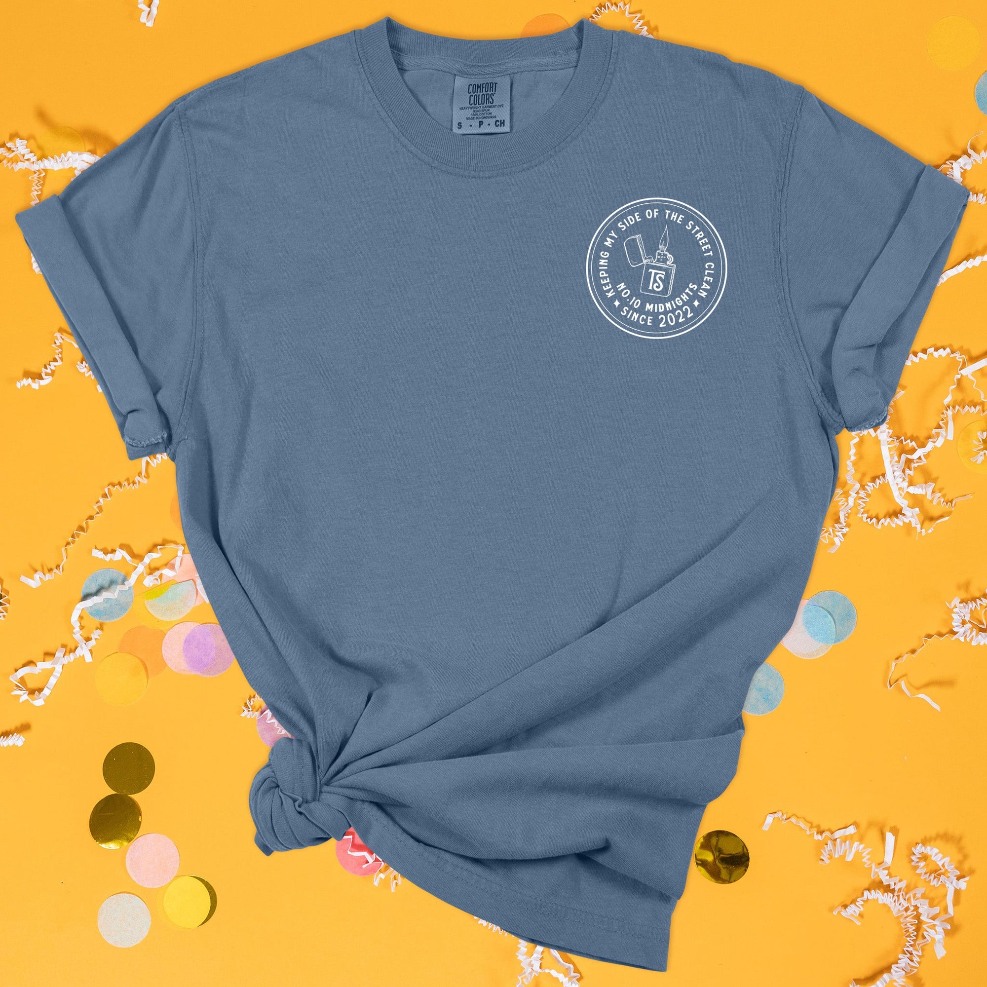 On a sunny mustard background sits the front of a t-shirt with white crinkle and big, colorful confetti scattered around. This Taylor Swift Inspired Reputation tee is dark blue with white lettering and a lighter over the left chest area. It says "KEEPING MY SIDE OF THE STREE CLEAN SINCE 2022" and "NO.10 MIDNIGHTS." The lighter has a "TS" on the front of it.