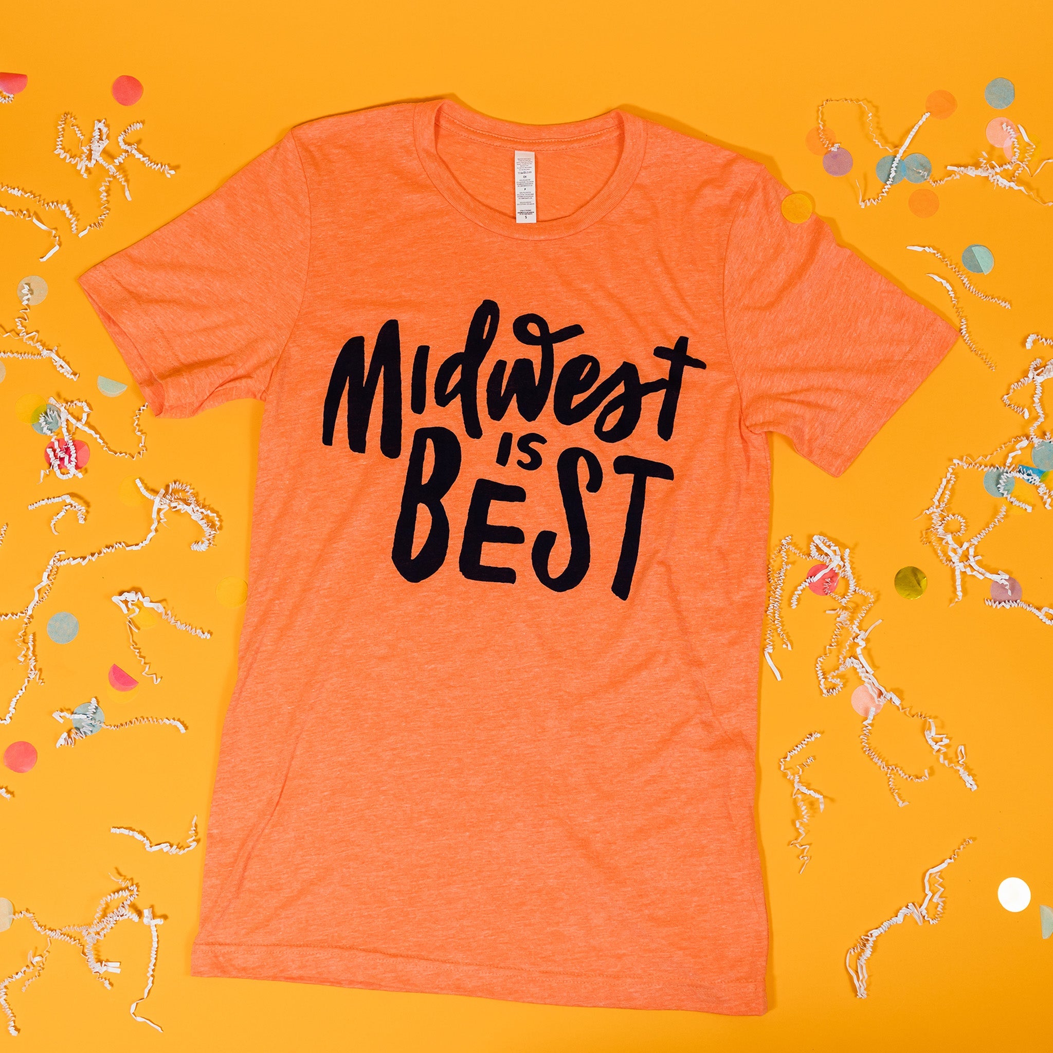 On a sunny mustard background sits a t-shirt with white crinkle and big, colorful confetti scattered around. This soft orange tee features custom hand lettering in navy ink and says "Midwest is BEST."