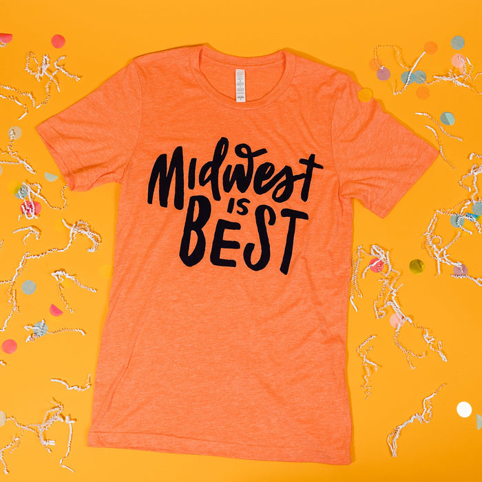 On a sunny mustard background sits a t-shirt with white crinkle and big, colorful confetti scattered around. This soft orange tee features custom hand lettering in navy ink and says "Midwest is BEST."