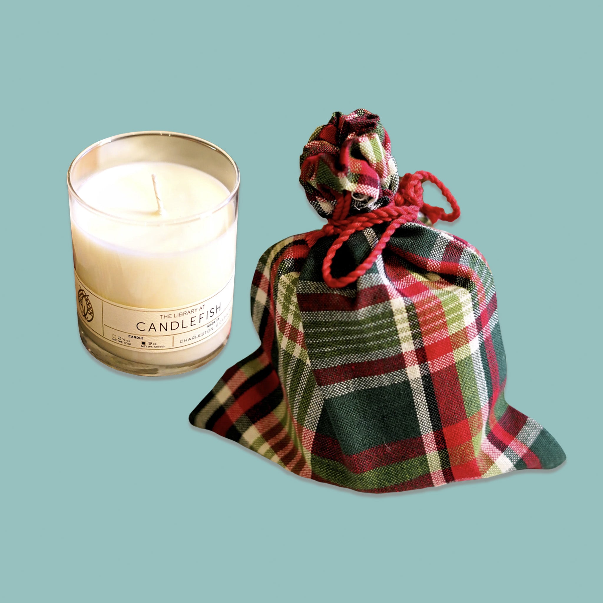 On a sage green background sits a classic library jar candle with a green plaid bag next to it. It is a '100% soy wax candle with a burn time of 15-20 hours for 2.5 ounces and 60-70 hours for 9 ounces'. 'Fragrance Notes: Eucalyptus, Fir Balsam, Woodland Pine. Fragrance Family - WOODS'.