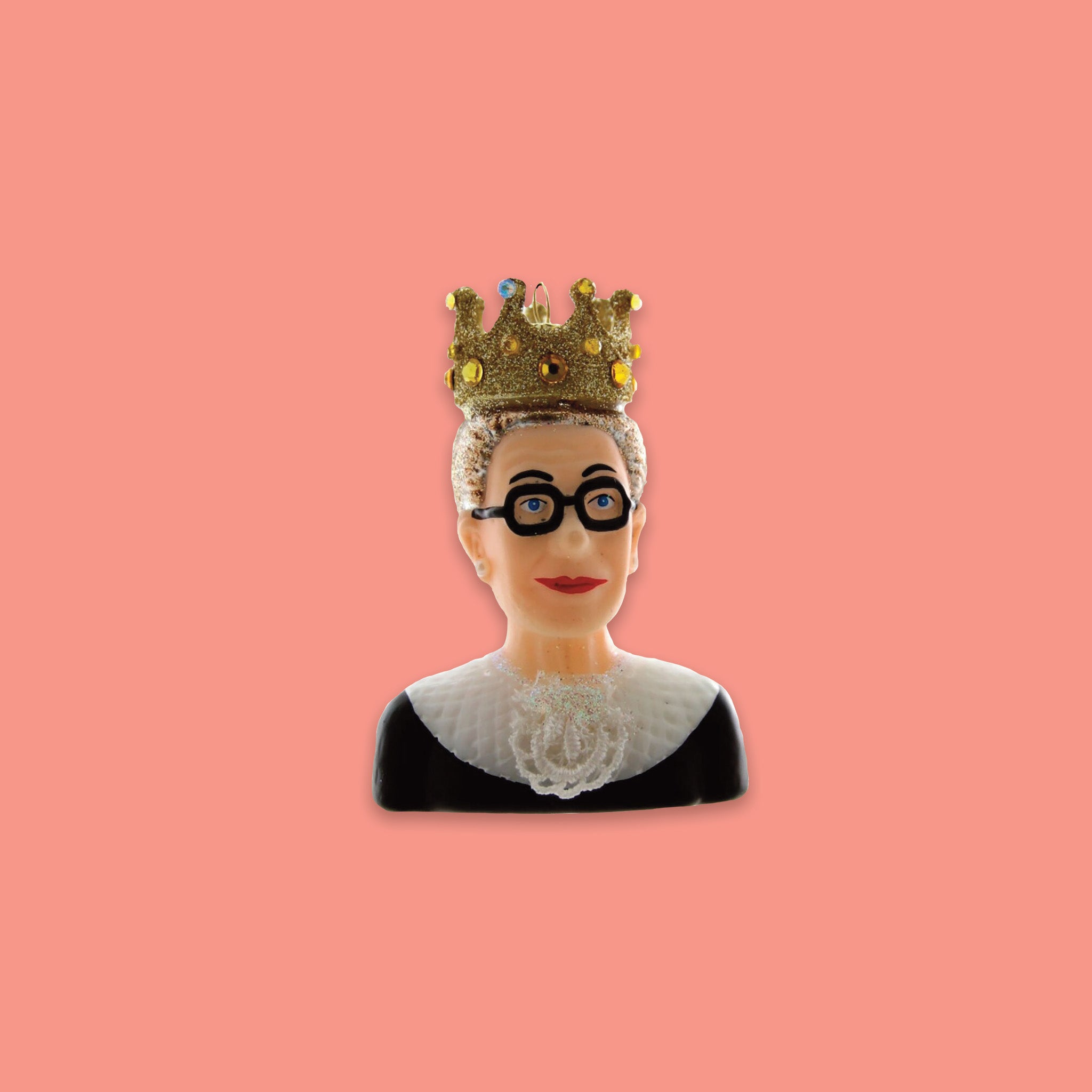 On a coral pink background sits an RBG inspired glass ornament. It is a bust of Ruth Bader Ginsberg wearing her black gown, her dissent collar, and a gold crown.