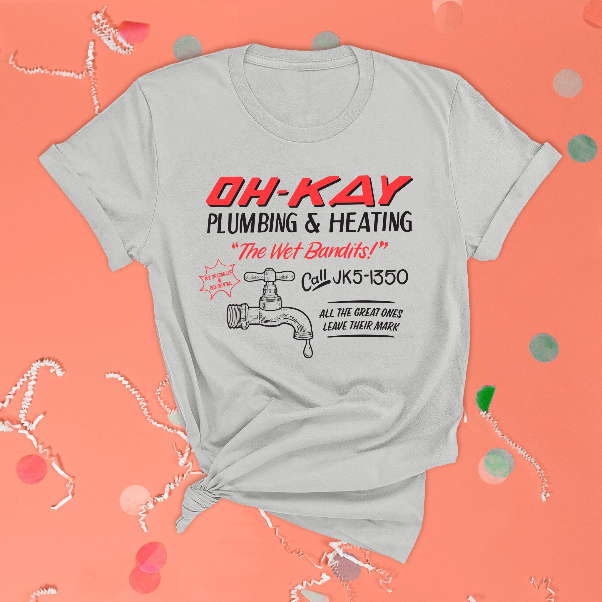 On a pink coral background sits the front of a t-shirt with white crinkle and big, colorful confetti scattered around. This Home Alone Inspired crewneck grey t-shirt has black and bright coral red lettering and an illustration of a water faucet. It says "OH-KAY PLUMBING & HEATING, "The Wet Bandits!", Call JK5-1350, ALL THE GREAT ONES LEAVE THEIR MARK, and WE SPECIALIZE IN RESIDENTIAL."