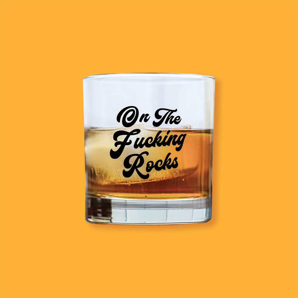 On a sunny mustard background is a glass. This low ball whiskey glass says "On The Fucking Rocks" in a black, handwritten lettering script font. It is filled with whiskey and ice.