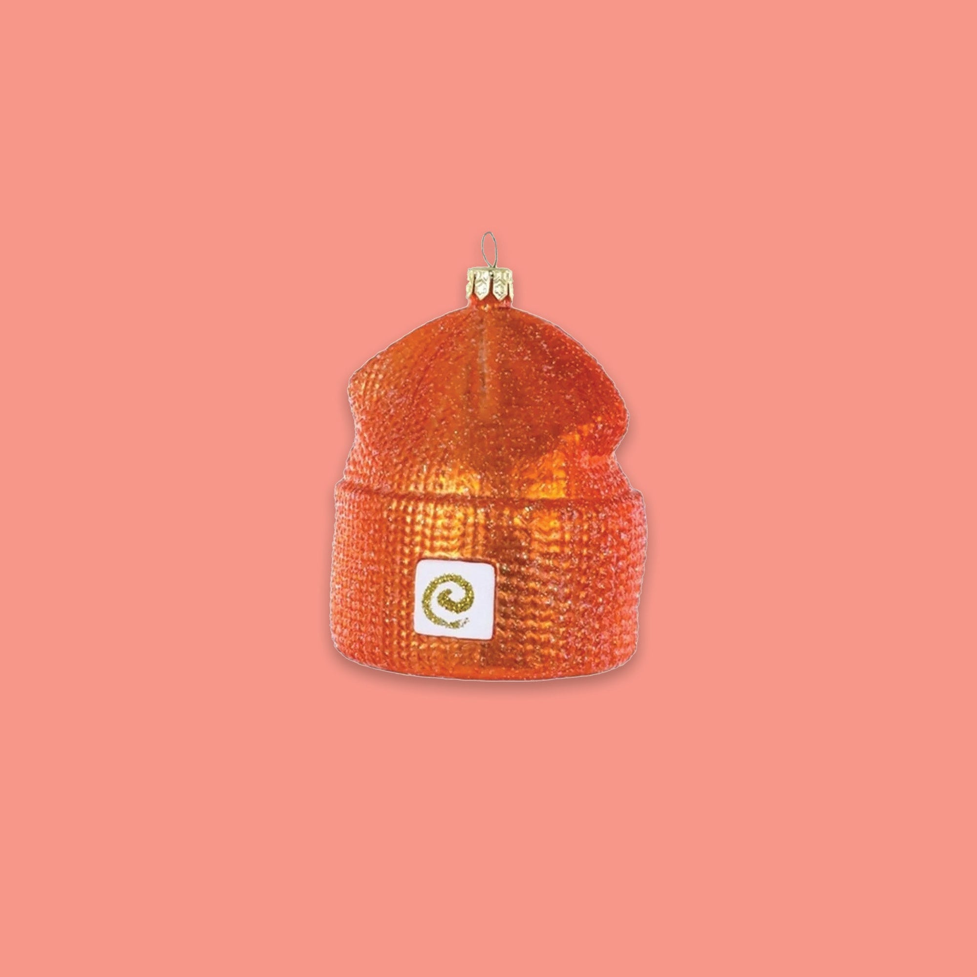 On a coral pink background sits a beanie ornament. This Carhartt inspired glass beanie ornament is orange.