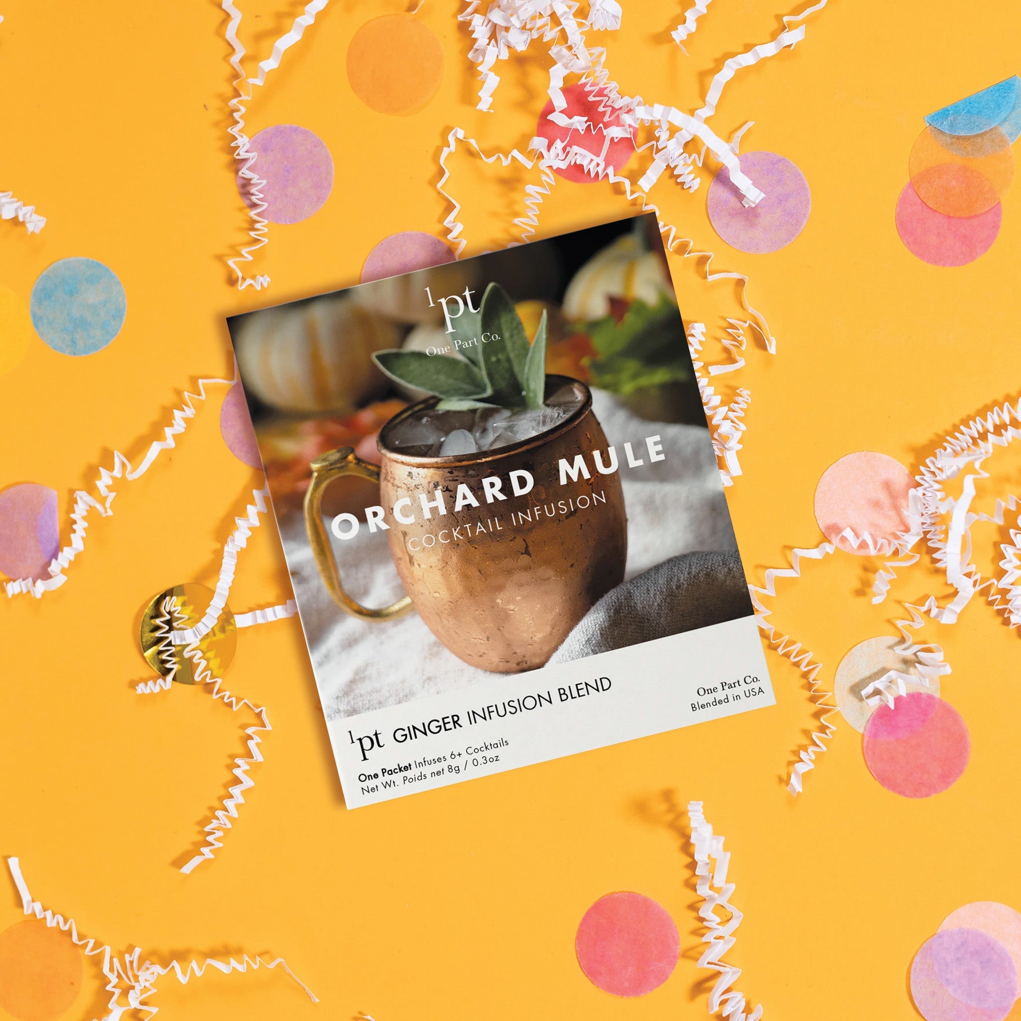 On a sunny mustard background sits a packet with a photo of an Orchard Mule in a copper mug with sage sprigs as garnish. It is an 'ORCHARD MULE COCKTAIL INFUSION' from One Part Co. One Packet infuses 6+ COCKTAILS. Net Wt. 8g / 0.3oz. There are white crinkle and big, colorful confetti scattered around.