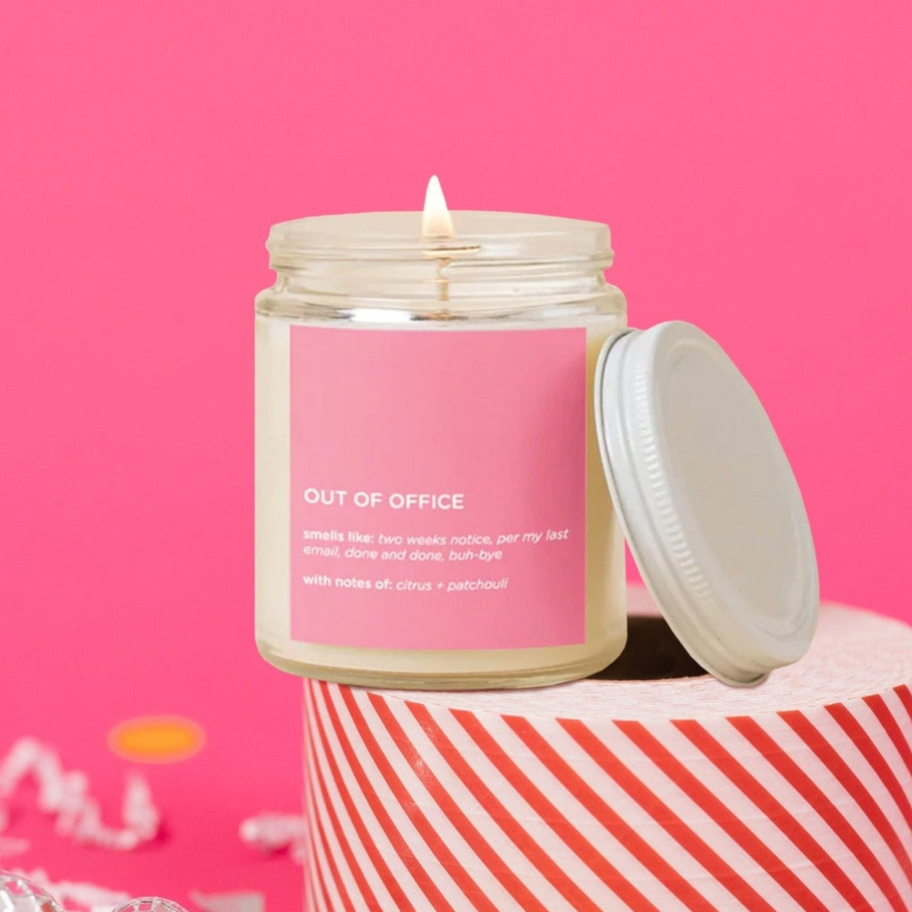 On a hot pink background sits candle and a lid. There are white crinkle and big, colorful confetti scattered around. The lit glass candle has a bubblegum pink label. It says "OUT OF OFFICE" in all caps, white block font. It says "smells like: two weeks notice, per my last email, done and done, buh-bye," and "with notes of: citrus + patchouli" in white, block font.