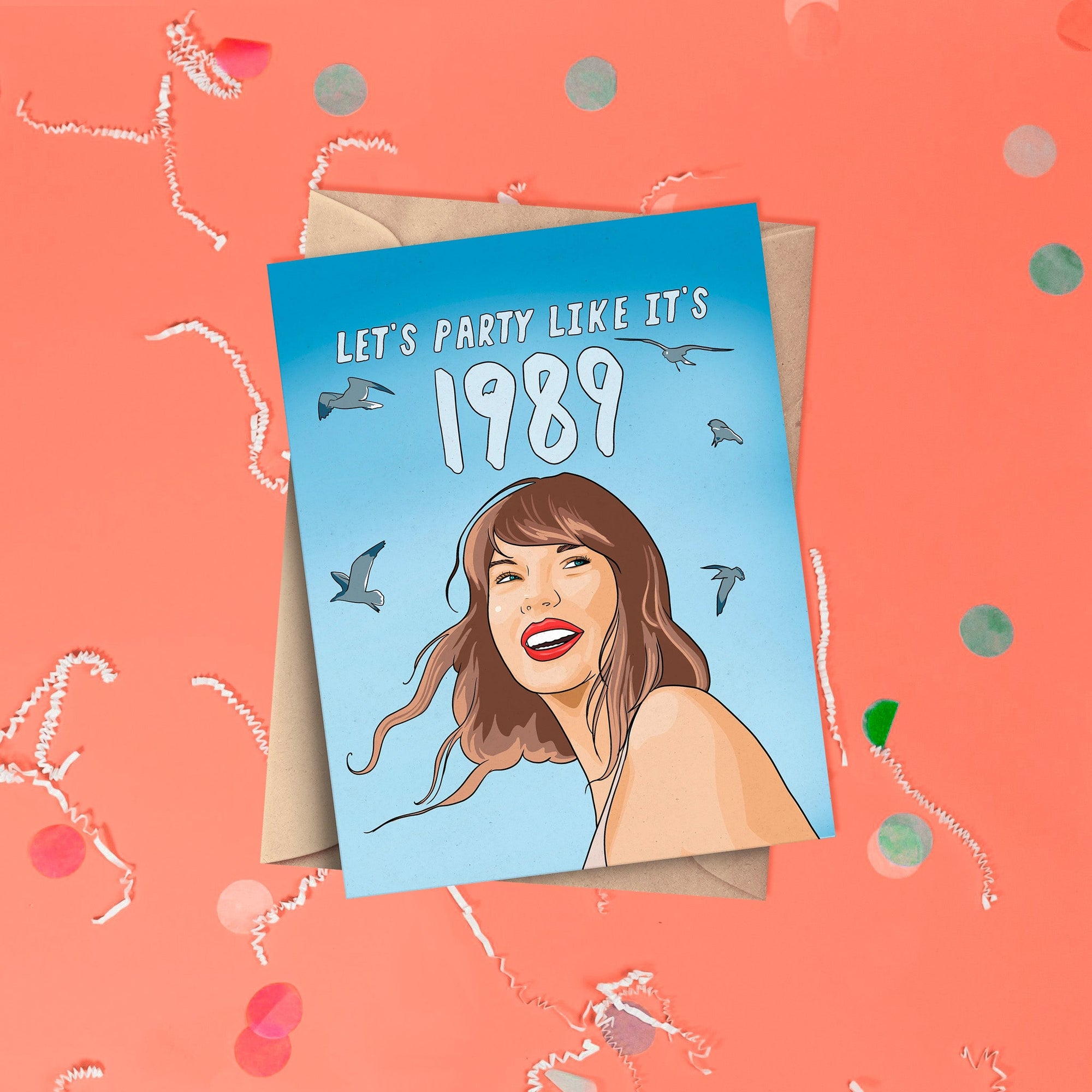 On a coral pink background sits a card with white crinkle and big, colorful confetti scattered around. This Taylor Swift inspired card has different shades of blue. There is an illustration of Taylor Swift and seagulls flying and it says "LET'S PARTY LIKE IT'S 1989" in white lettering. It has a kraft envelope.