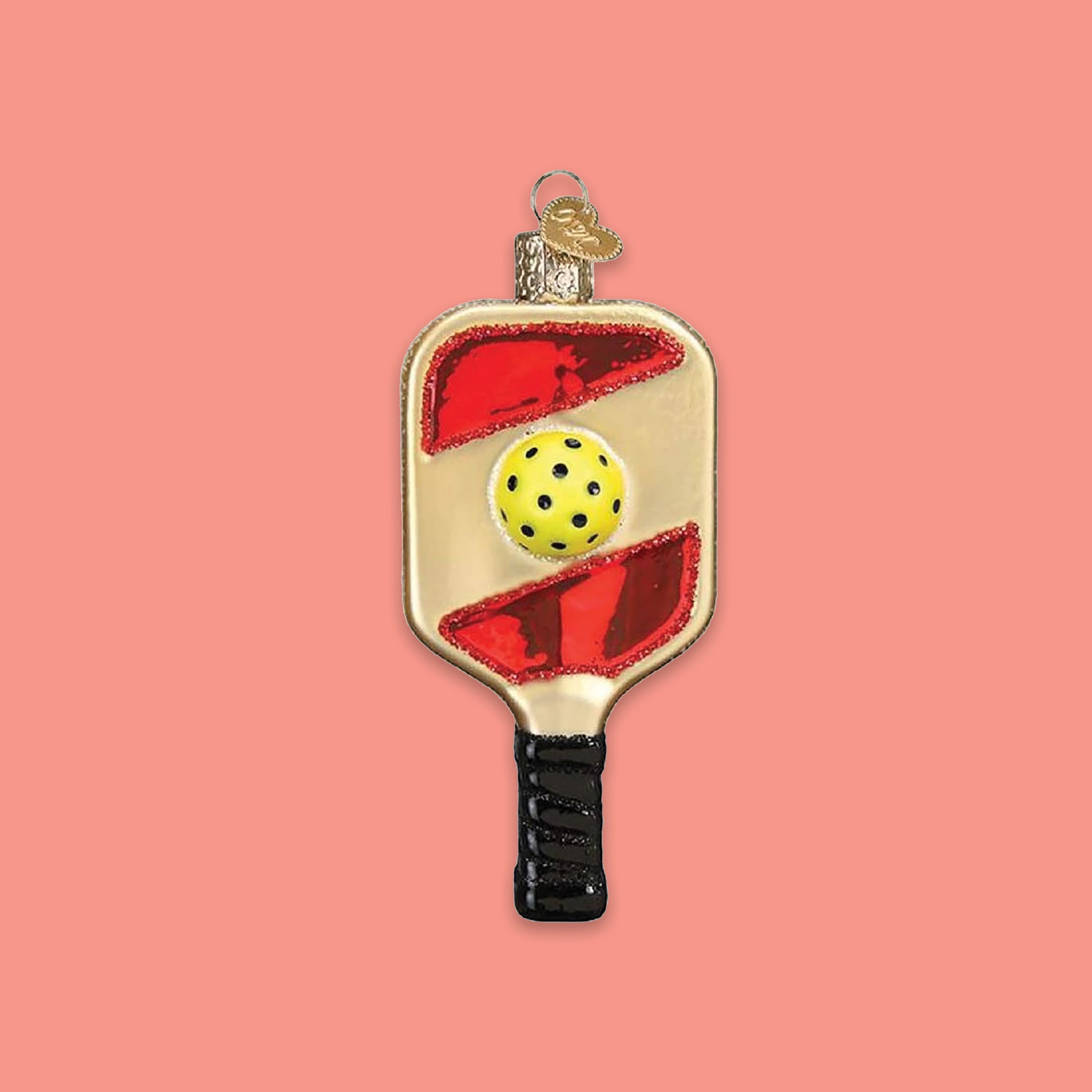 On a coral pink background sits an ornament. This glass ornament is a red and gold pickleball paddle with a yellow ball.