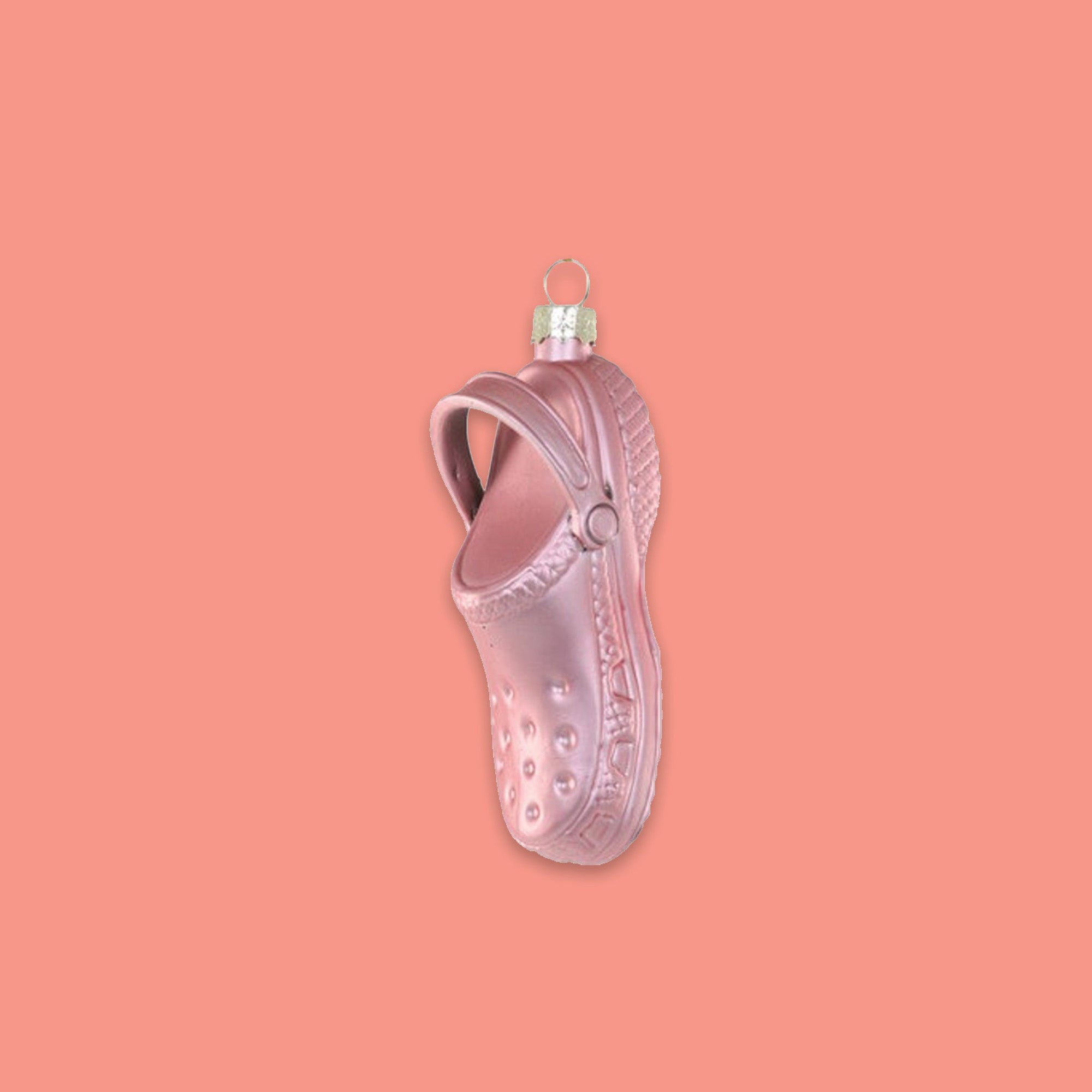 On a coral pink background sits an ornament. This glass ornament is a pink croc shoe.