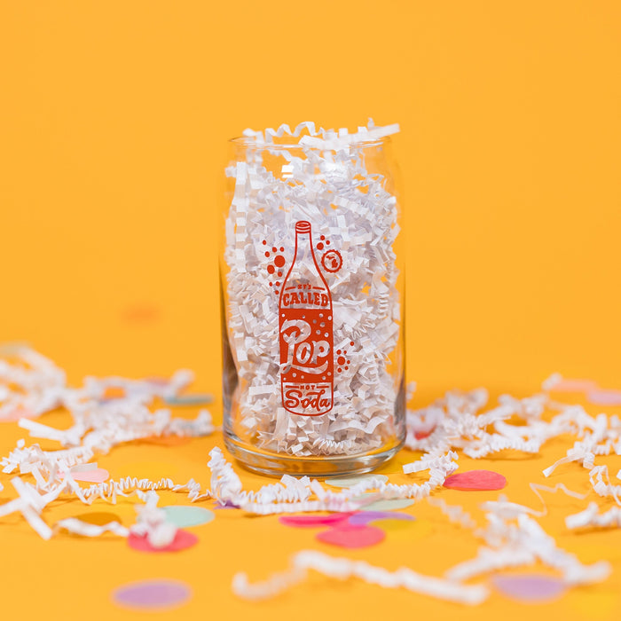 On a sunny mustard background sits a pint glass with white crinkle and big, colorful confetti scattered around. This beer can-style glass has a red and white handdrawn soda pop that says "It's Called Pop Not Soda" in red and white handwritten lettering. It has a white outline bubble with the state of Michigan in it in white and it is filled with white crinkle. 