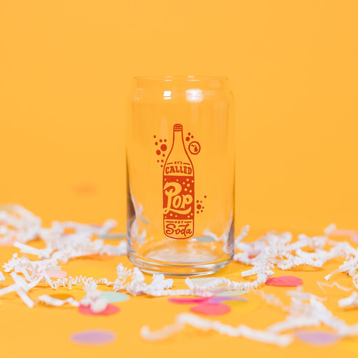 On a sunny mustard background sits a pint glass with white crinkle and big, colorful confetti scattered around. This beer can-style glass has a red and white handdrawn soda pop that says "It's Called Pop Not Soda" in red and white handwritten lettering. It has a white outline bubble with the state of Michigan in it in white.