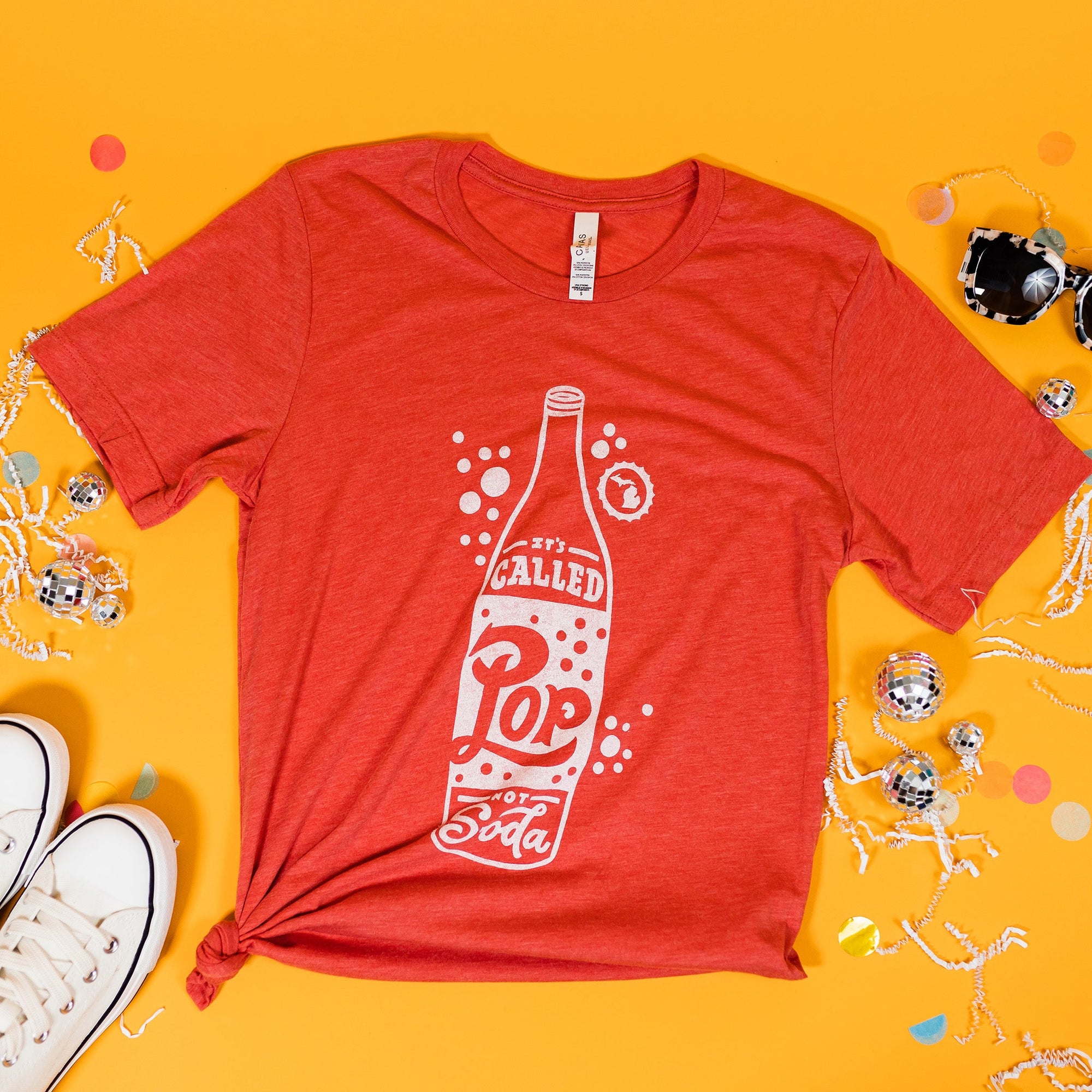 On a sunny mustard background sits a t-shirt with white crinkle and big, colorful confetti scattered around. There are mini disco balls, sunglasses, and a pair of white sneakers. This heather red tee features a custom illustration of a bottle of pop with bubbles and in white hand-lettering, it says "IT'S CALLED Pop NOT Soda."
